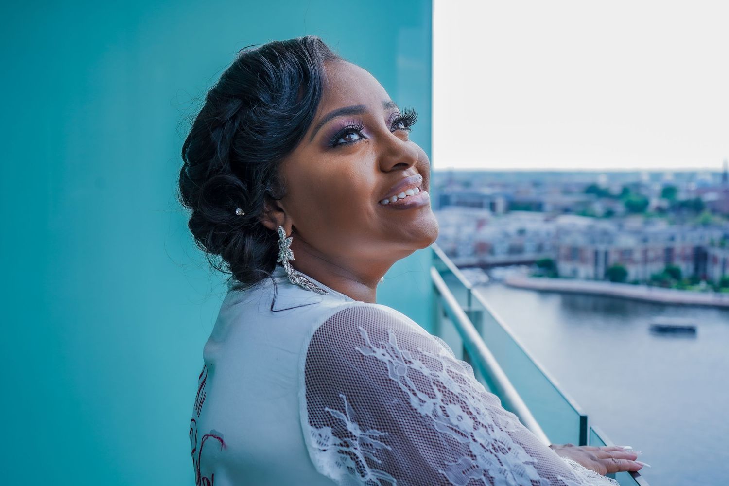 Bride in elegant lace dress poses by window with city skyline view.