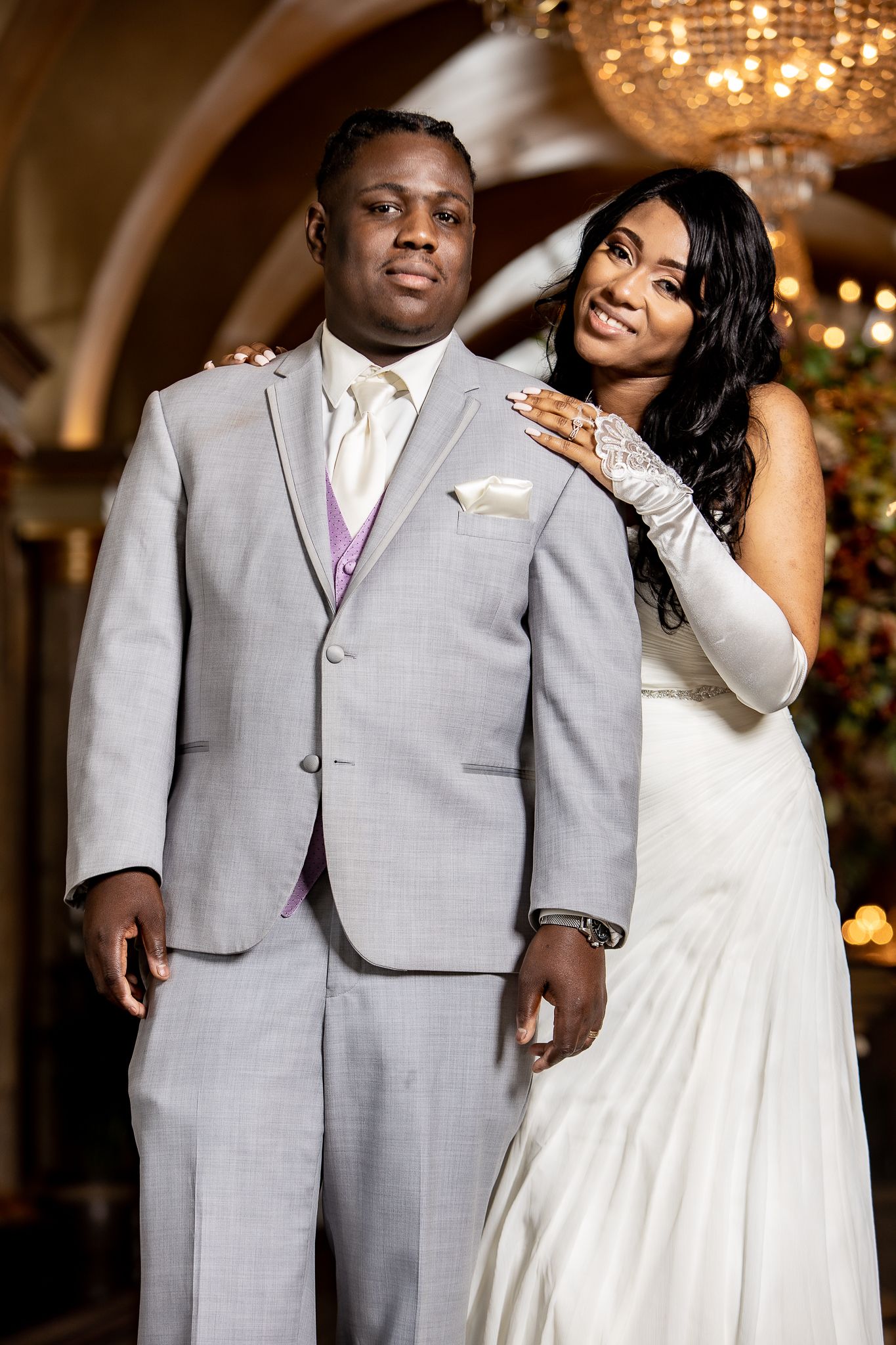 Couple in wedding attire pose beneath ornate chandeliers in elegant venue hallway.