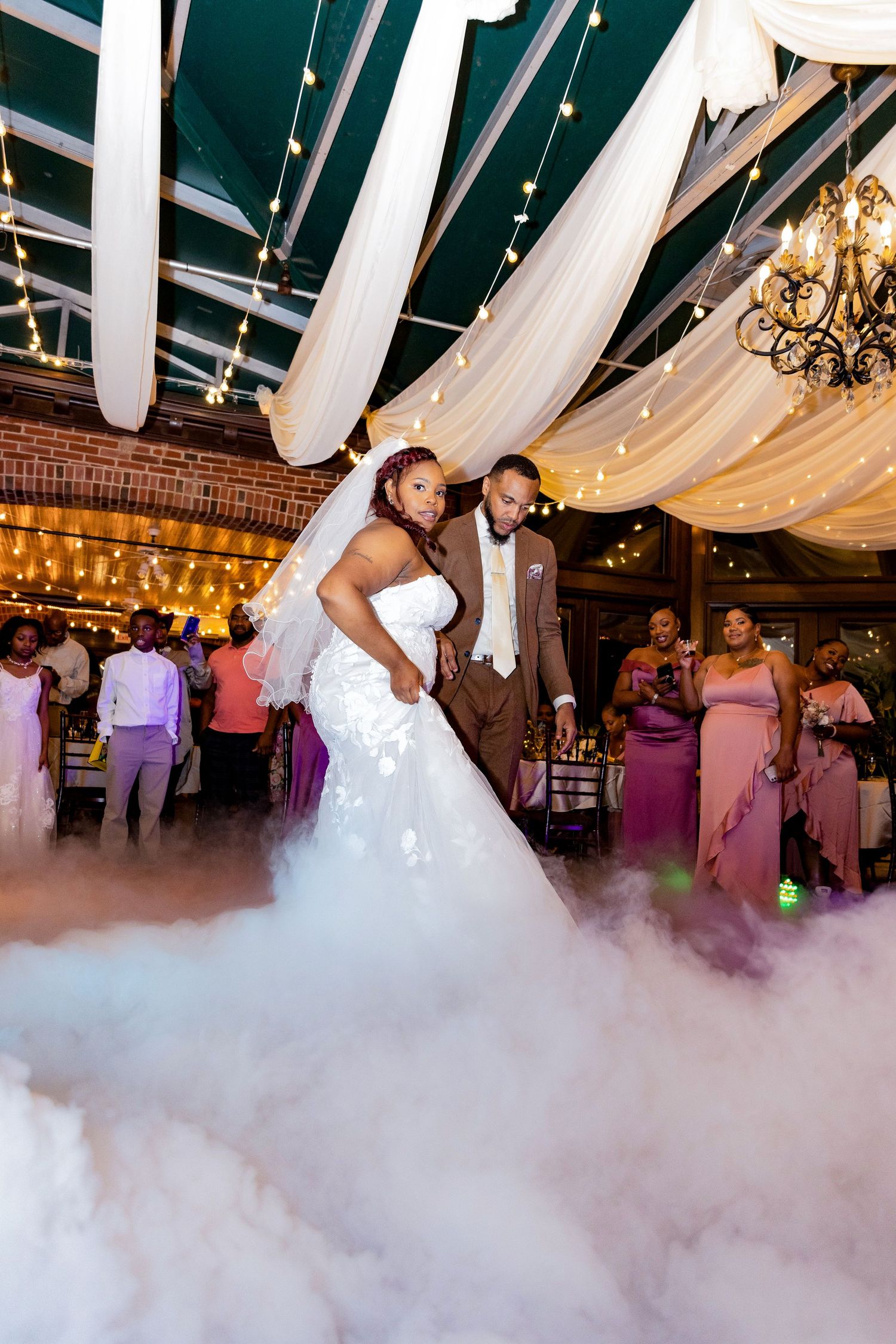 A couple dances amid fog effects under draped fabric and chandeliers at their reception.