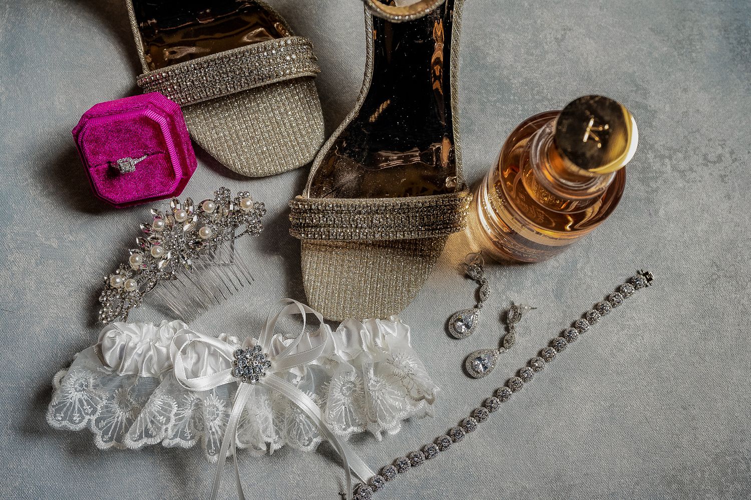 Wedding accessories including garter, perfume bottle, and jewelry arranged on grey surface.