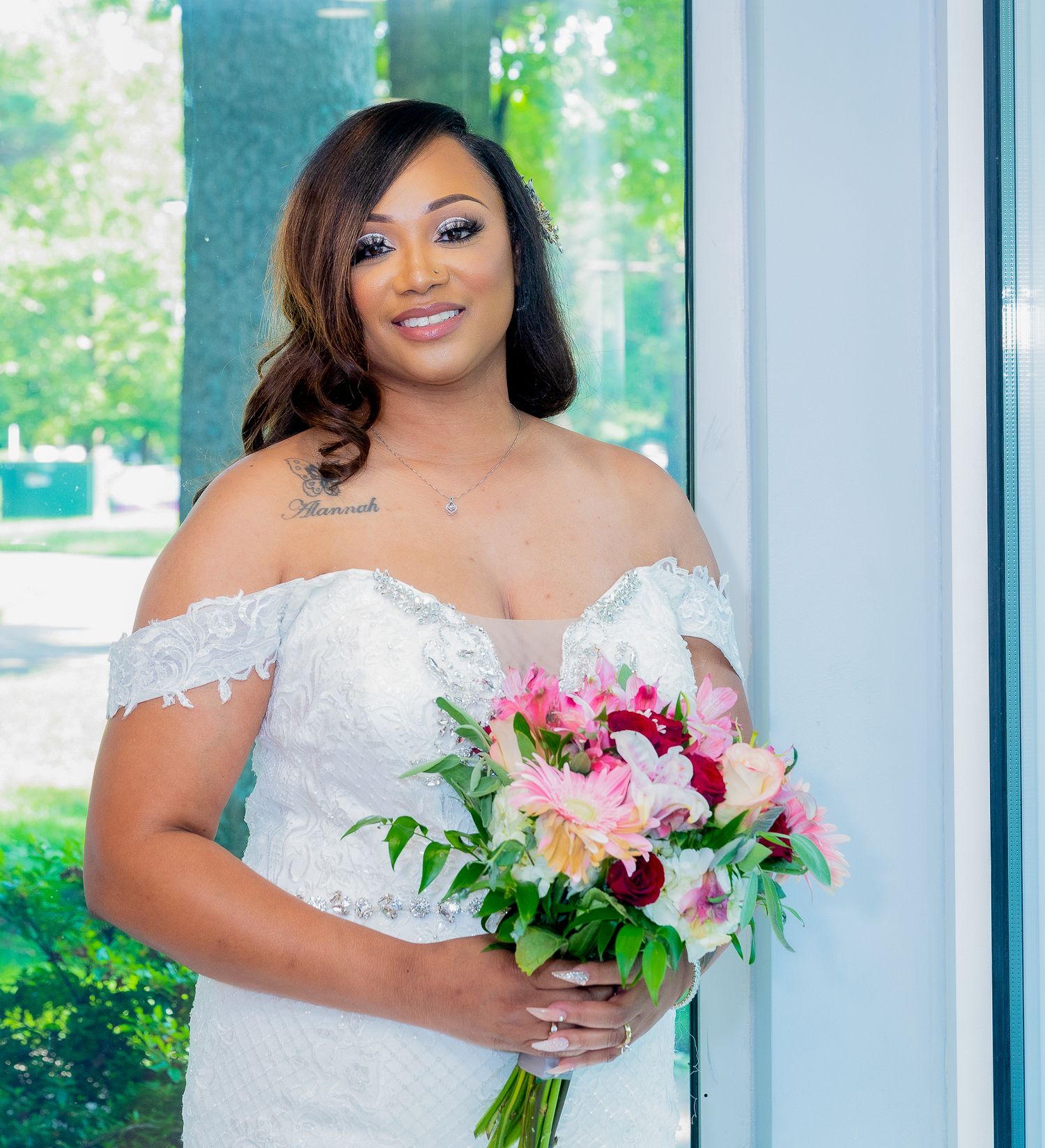 Beautiful bride in a white lace dress holding a pink and white floral bouquet against a bright window.