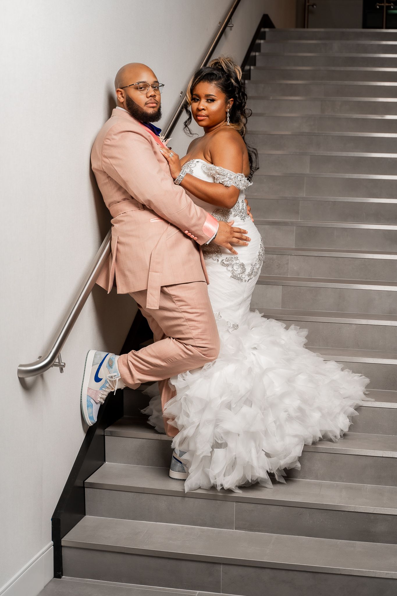 Stylish couple posing on stairs with bride in ruffled gown and groom in pink suit with sneakers.