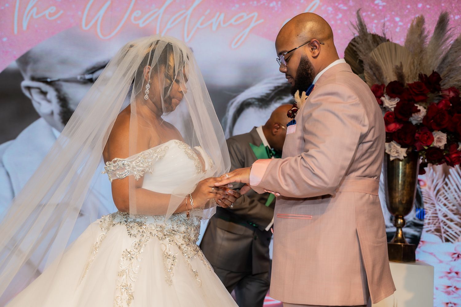 A wedding ceremony moment with a couple exchanging rings in front of a pink backdrop.
