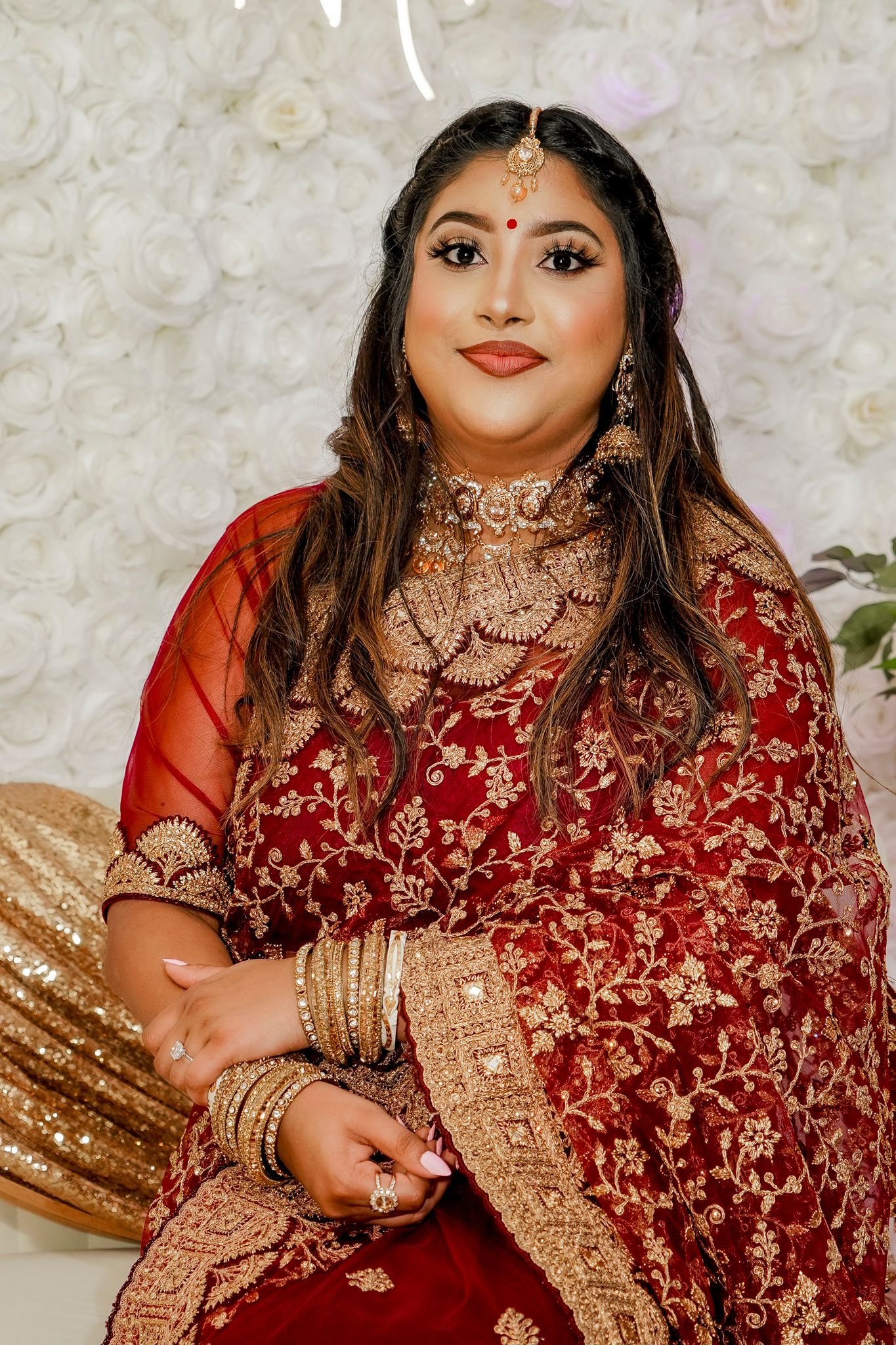 Woman in traditional red and gold Indian bridal attire poses against floral backdrop.