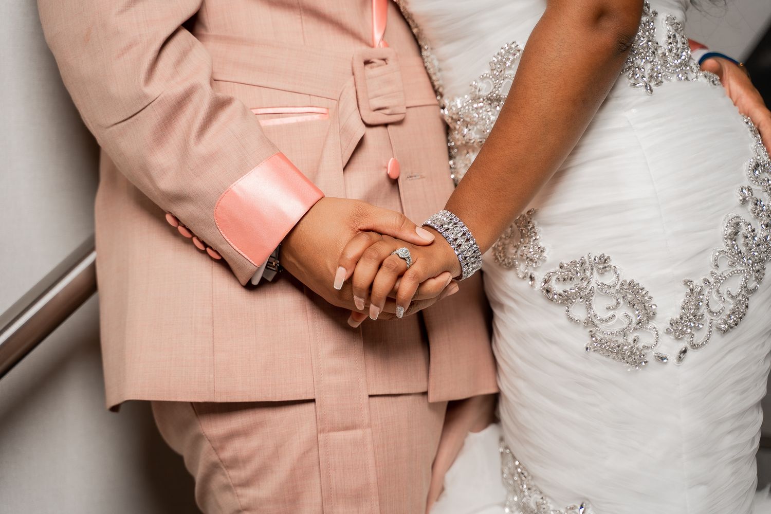 Close-up detail of newlyweds holding hands, showing blush suit and white lace dress.