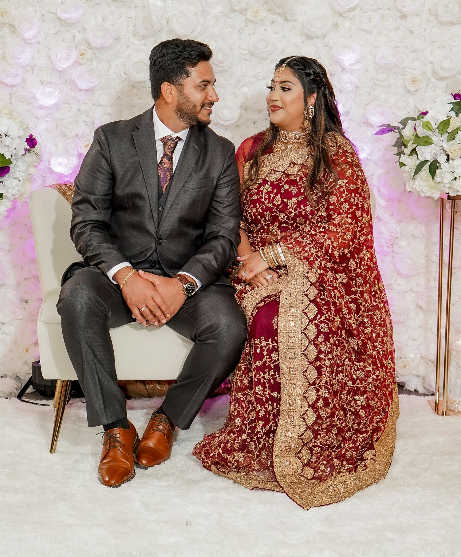 A couple in traditional Indian wedding attire posing together against a floral backdrop.