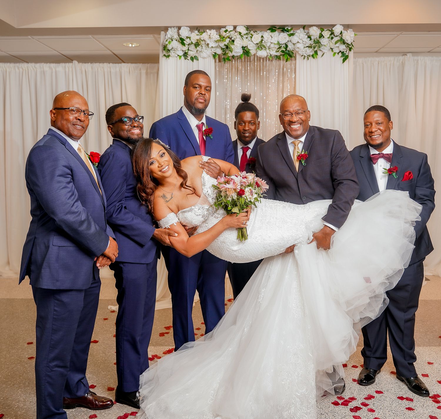 Wedding party in navy suits and white dress posing in front of floral backdrop.