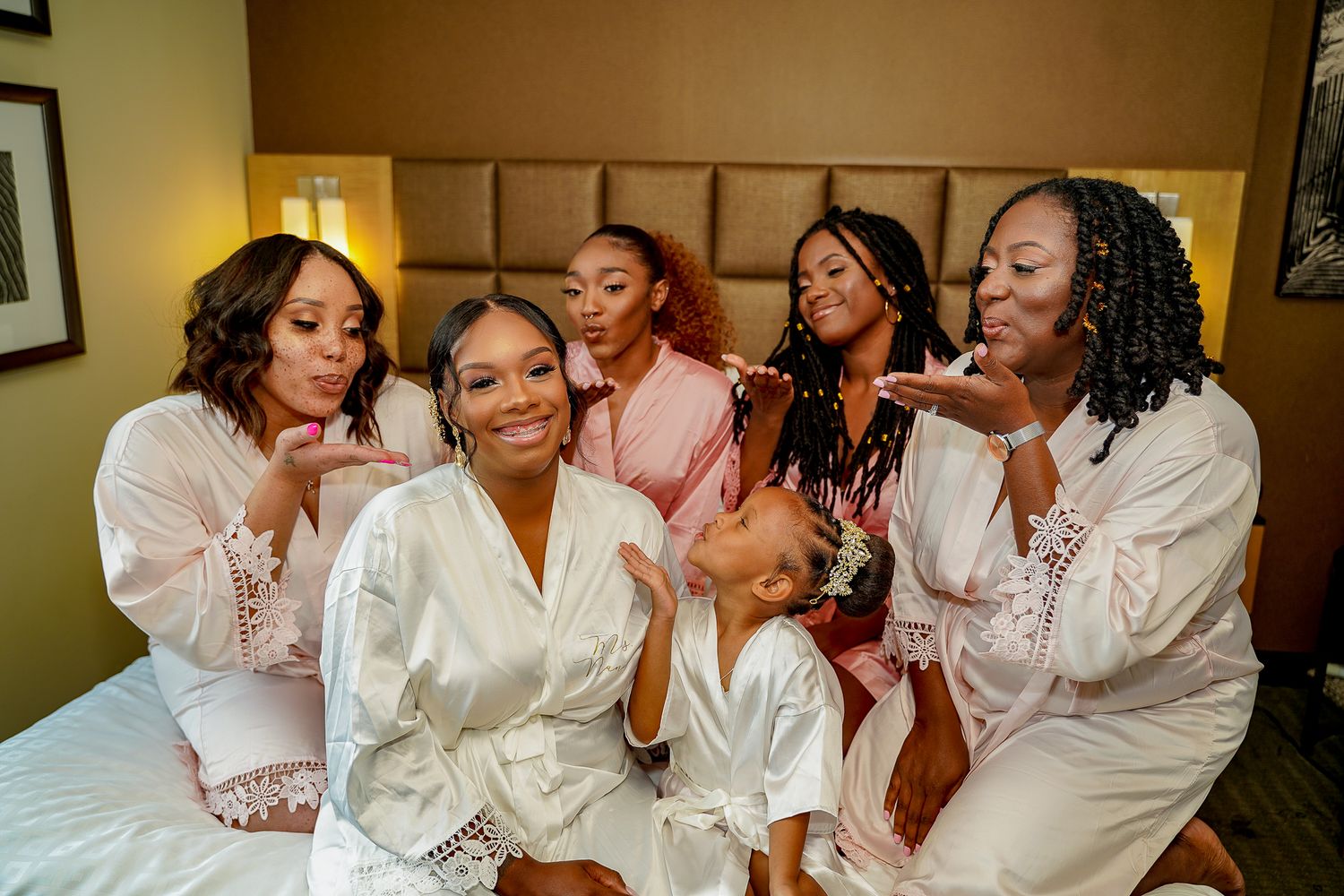 Group of bridesmaids in white silk robes laughing and embracing during pre-wedding preparations.