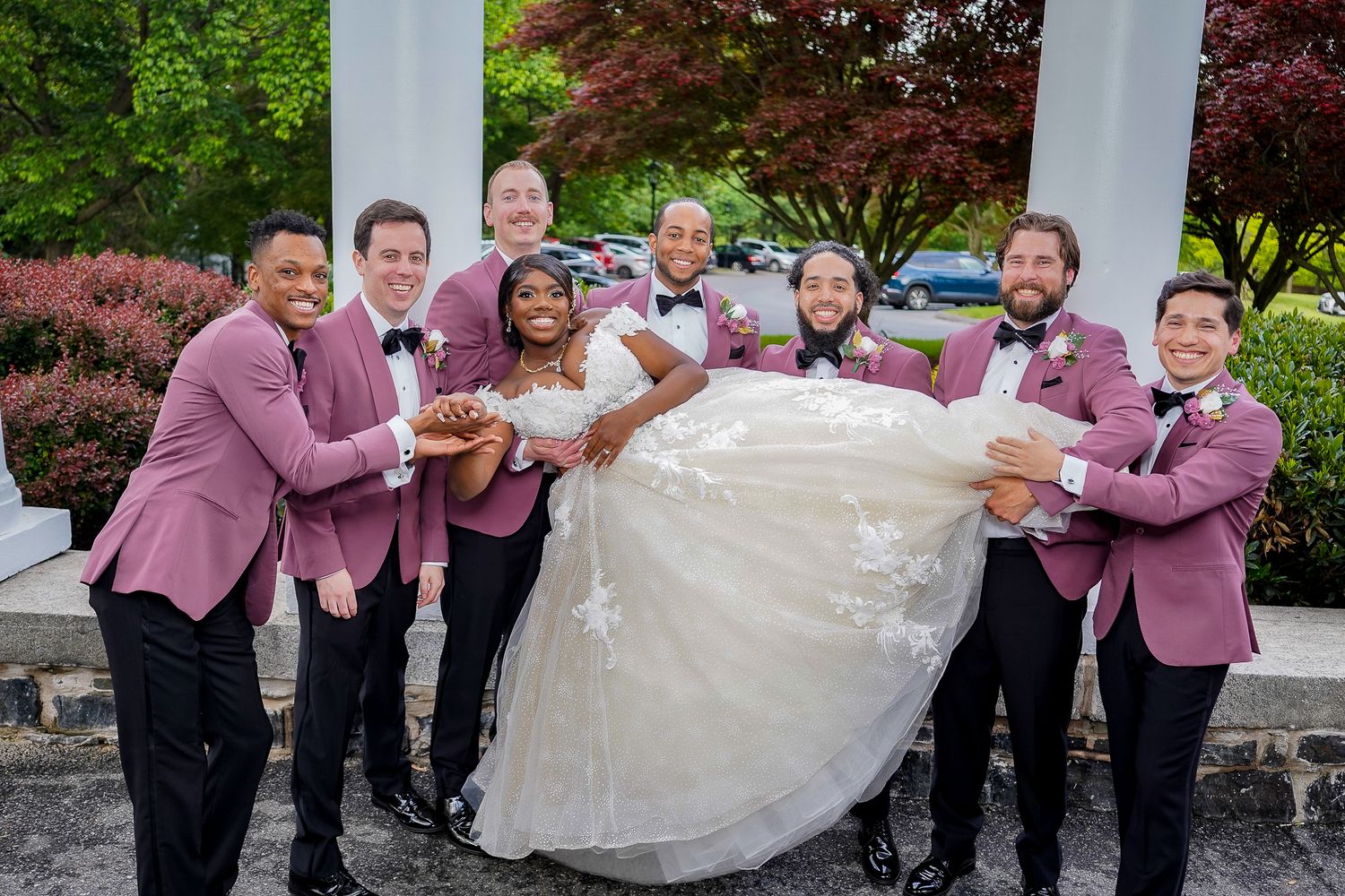 Wedding party members lift bride in white dress during fun group photo.