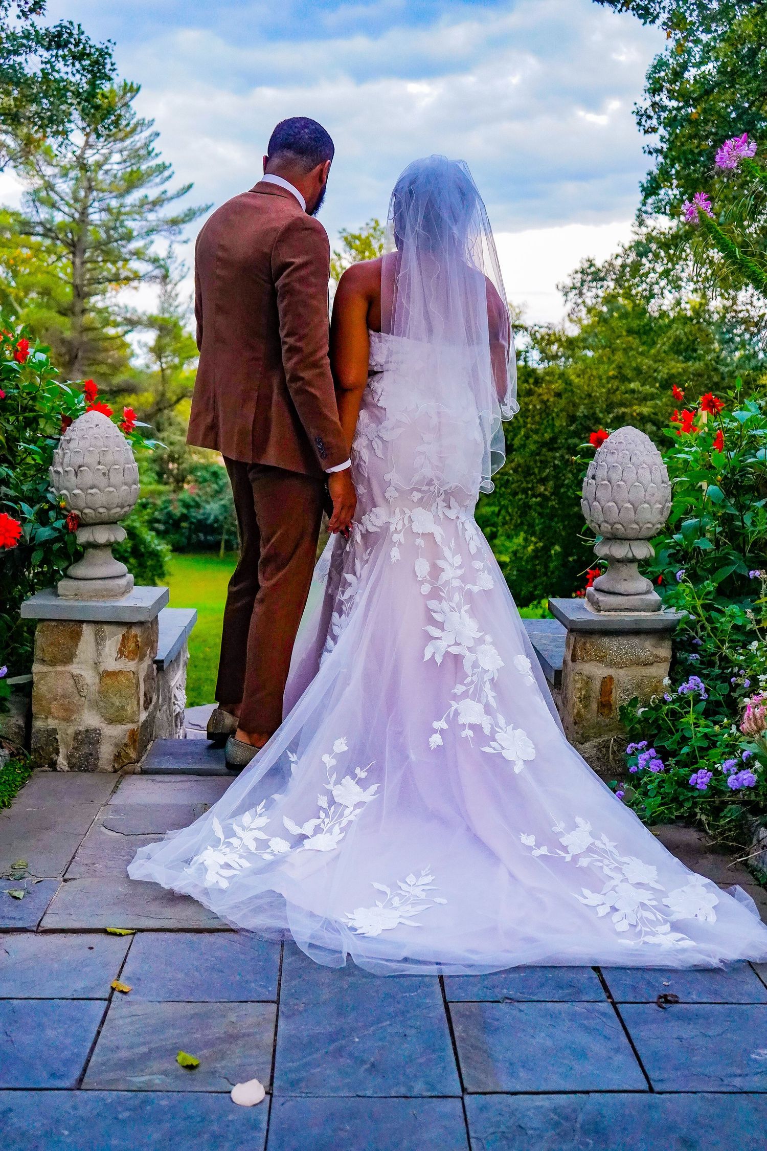 Bride in flowing white gown and groom in brown suit pose on stone patio surrounded by garden flowers.