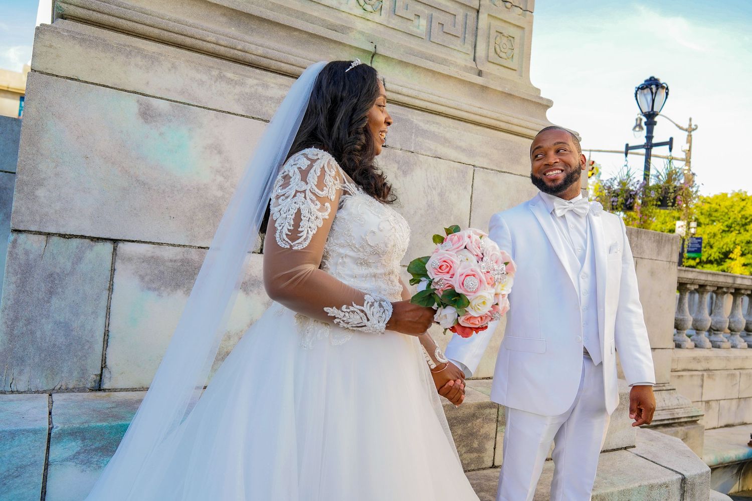 A couple in white wedding attire poses near a stone architectural feature.