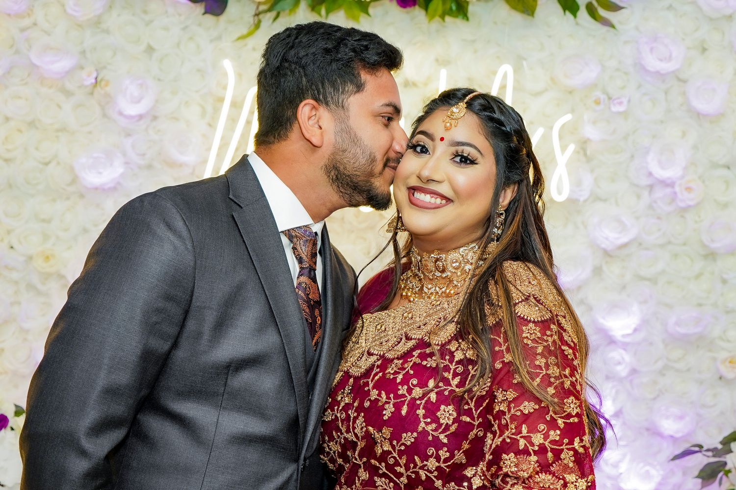 Wedding couple in traditional red and black attire posing against an elegant white floral backdrop.