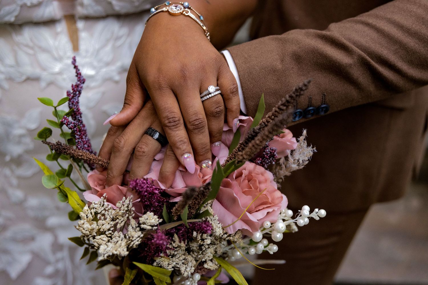 Close-up of wedding rings and hands holding a beautiful pink and purple floral bouquet.