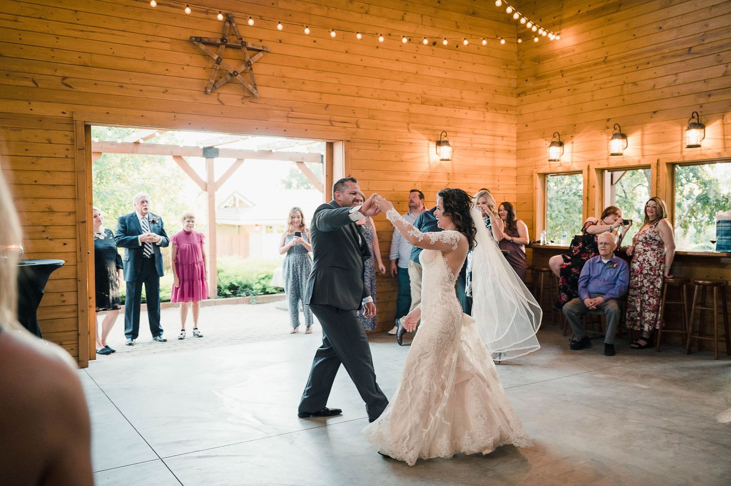 Wedding couple shares their first dance in a rustic wooden barn venue with string lights and guests watching.