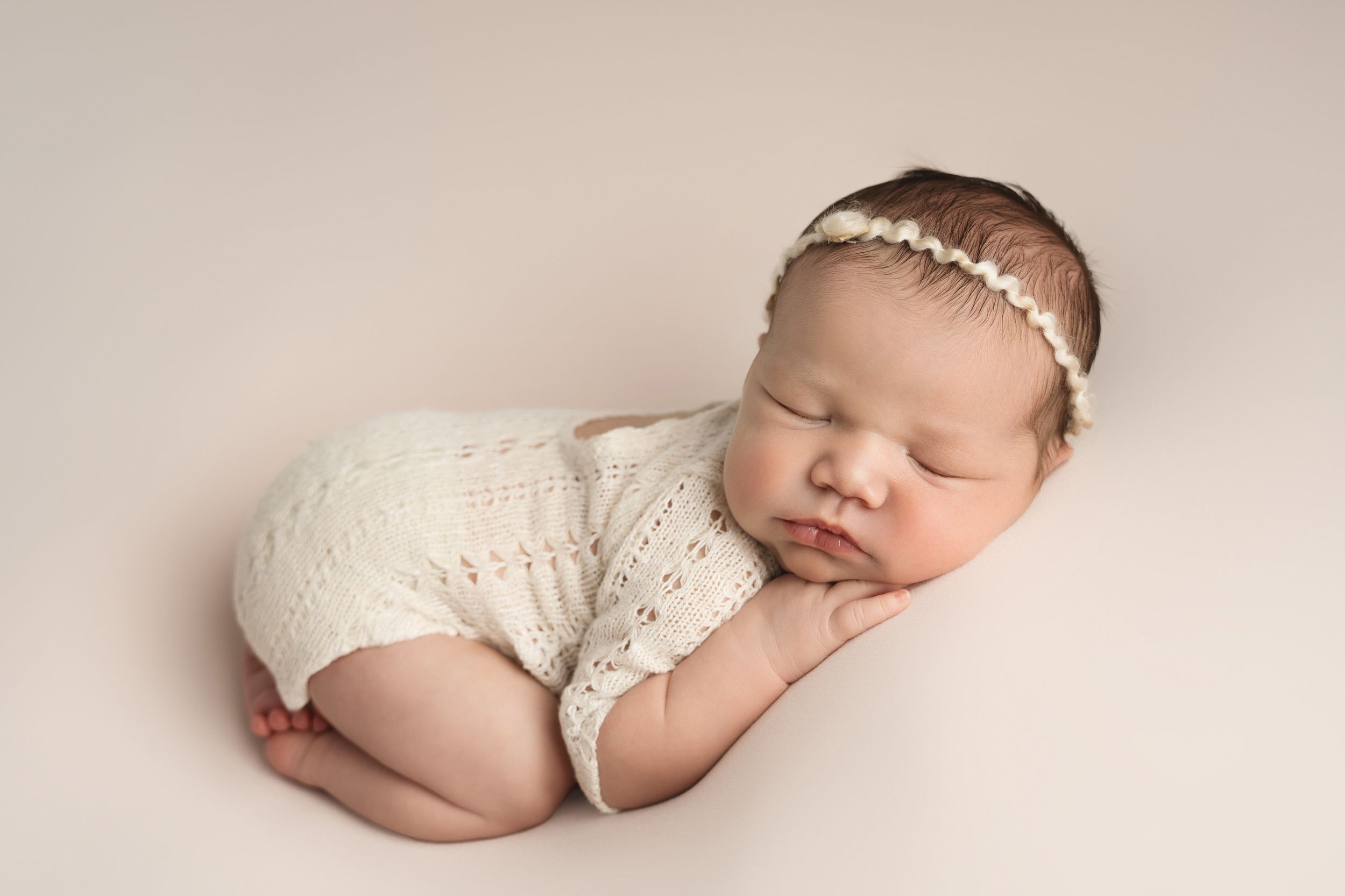 Peaceful newborn baby girl wearing white knit outfit and floral headband sleeping on soft pink backdrop.