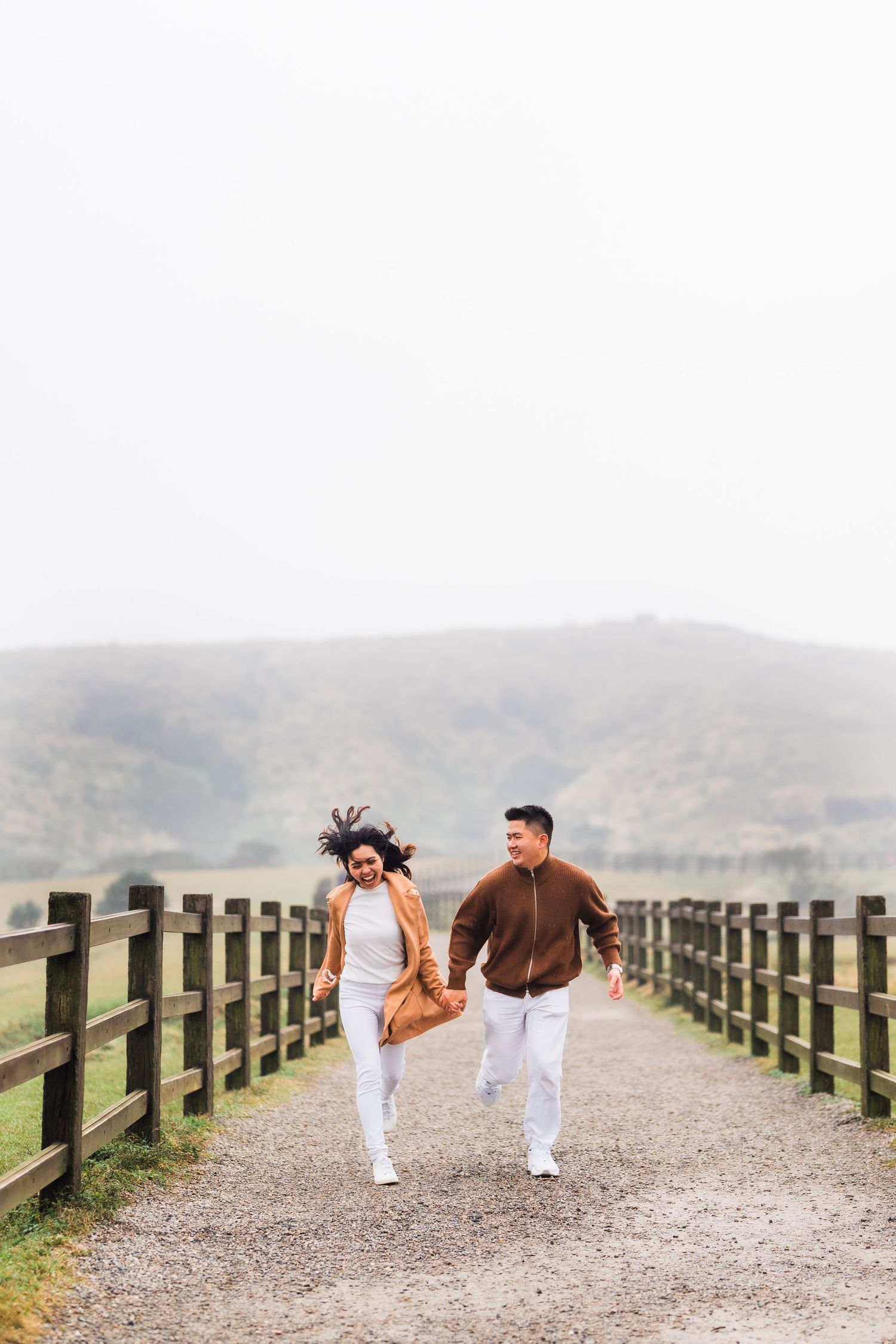 Couple running playfully on dirt path between wooden fences with mountains in background.