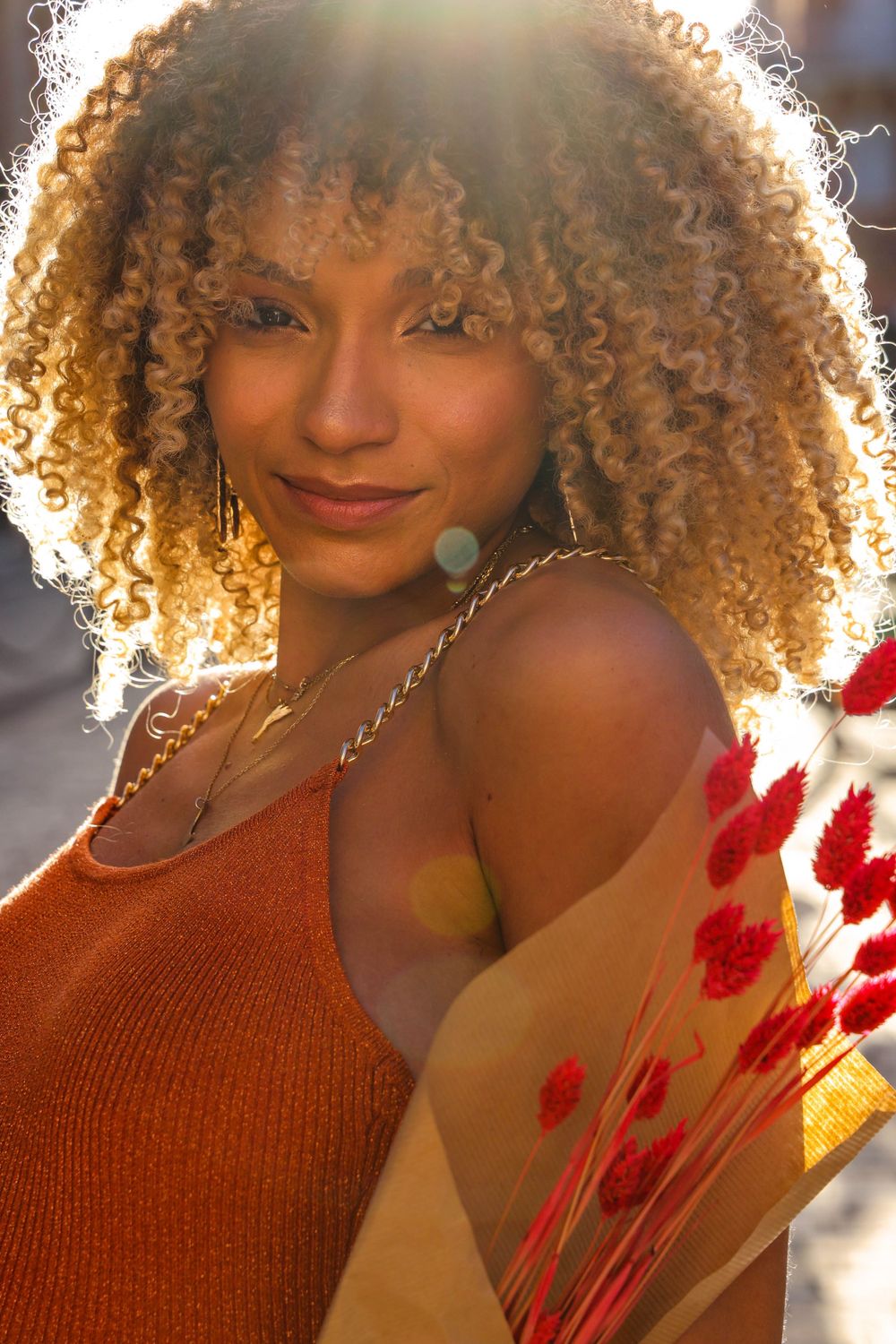Woman with curly blonde hair smiling in orange dress holding dried flowers in golden sunlight.