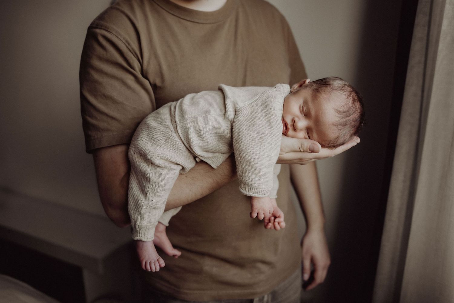 A drowsy baby in white clothing rests across someone wearing a brown sweater.
