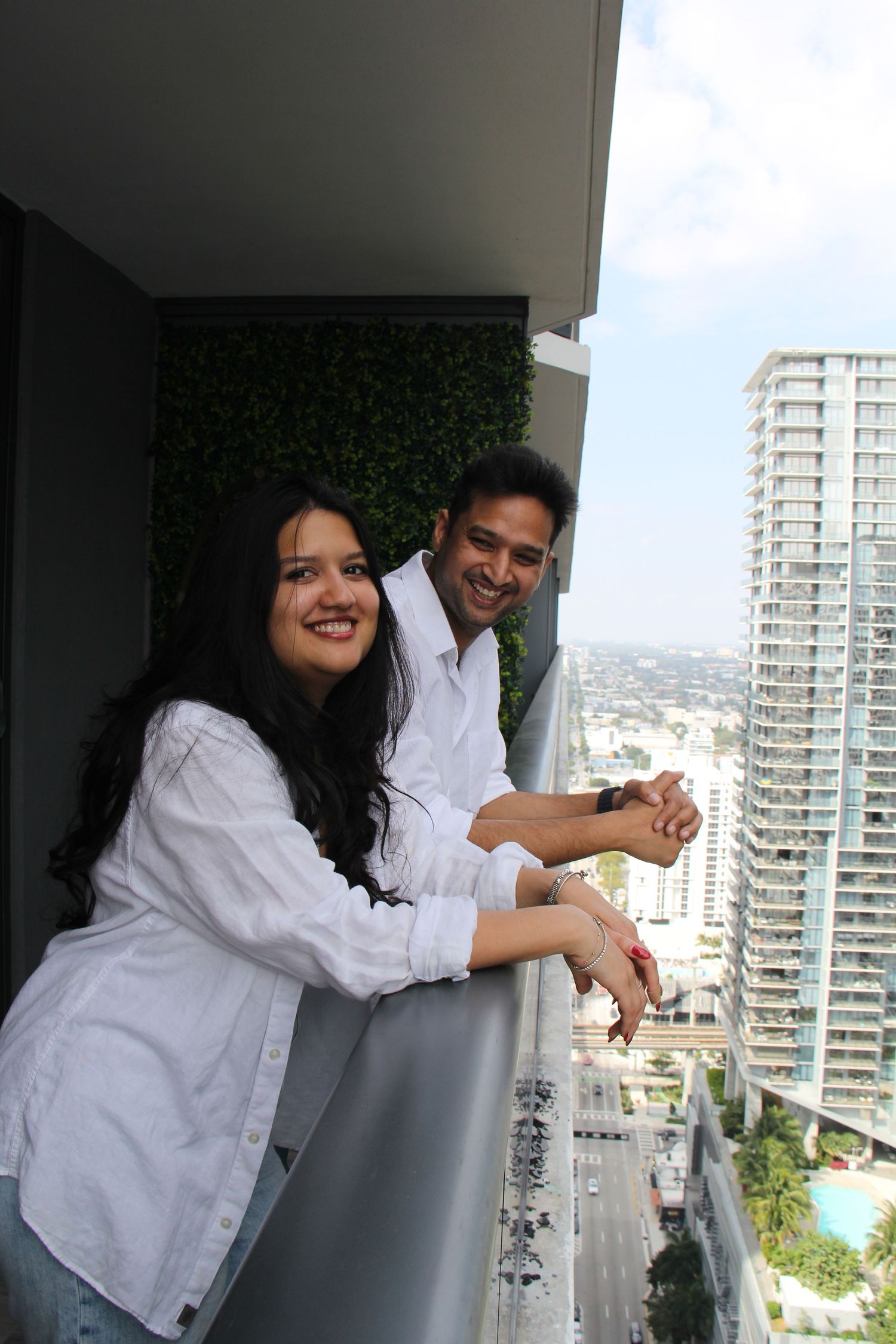 Two people in white shirts lean against a balcony railing overlooking a city skyline.