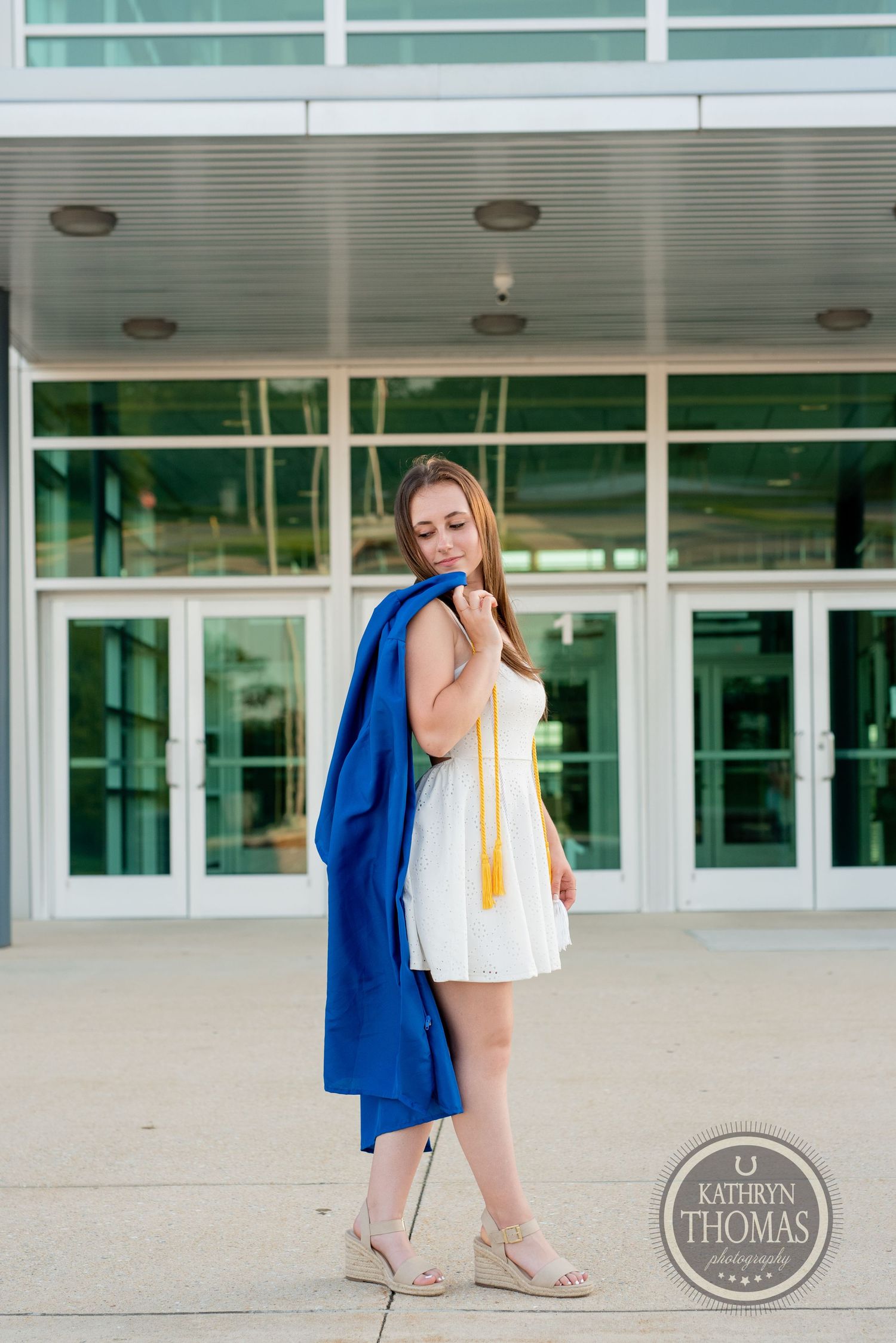 Graduate carries blue graduation gown while wearing white dress outside school building.