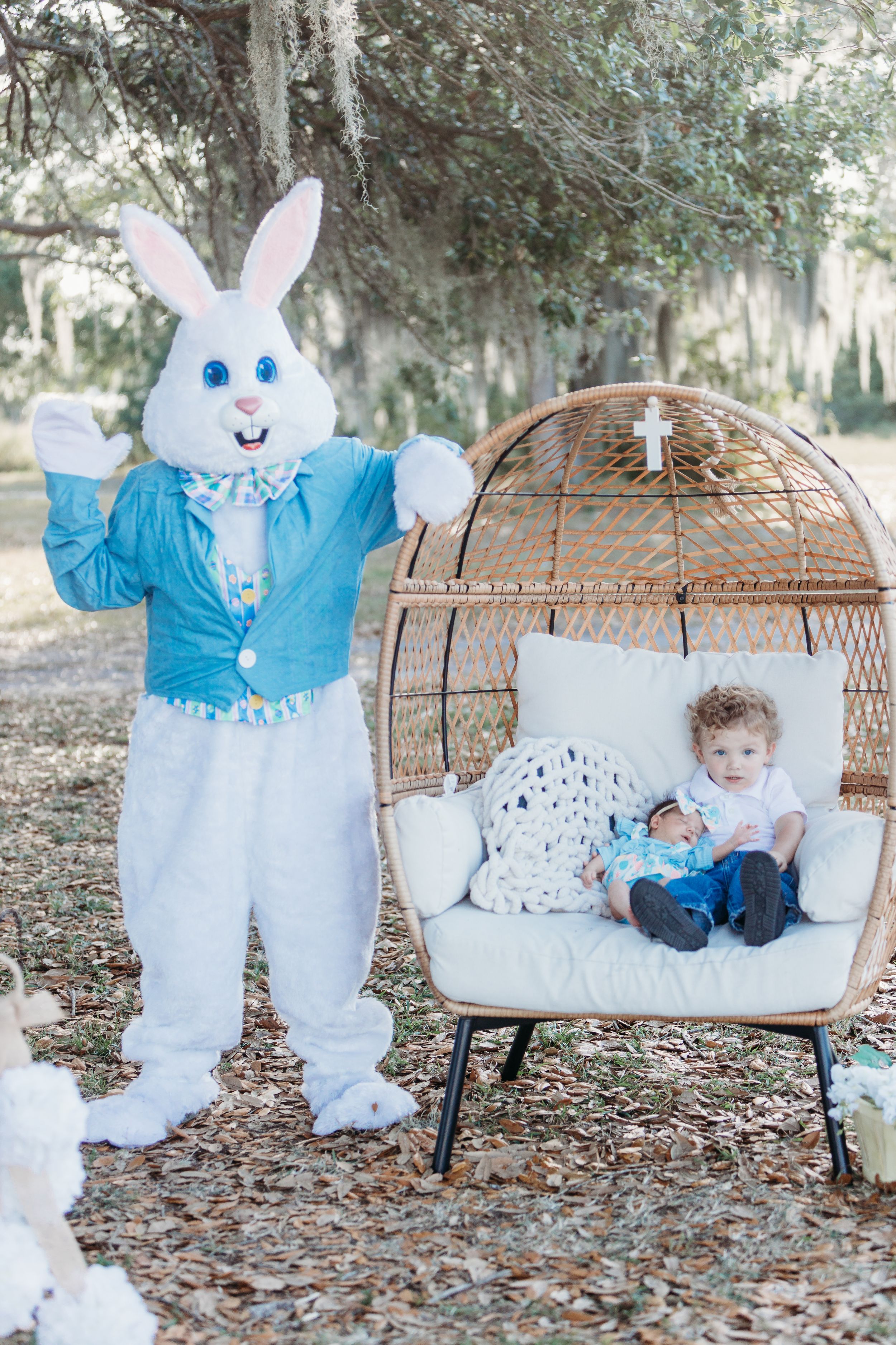 White Easter bunny mascot in blue jacket stands beside an outdoor wicker chair under trees.
