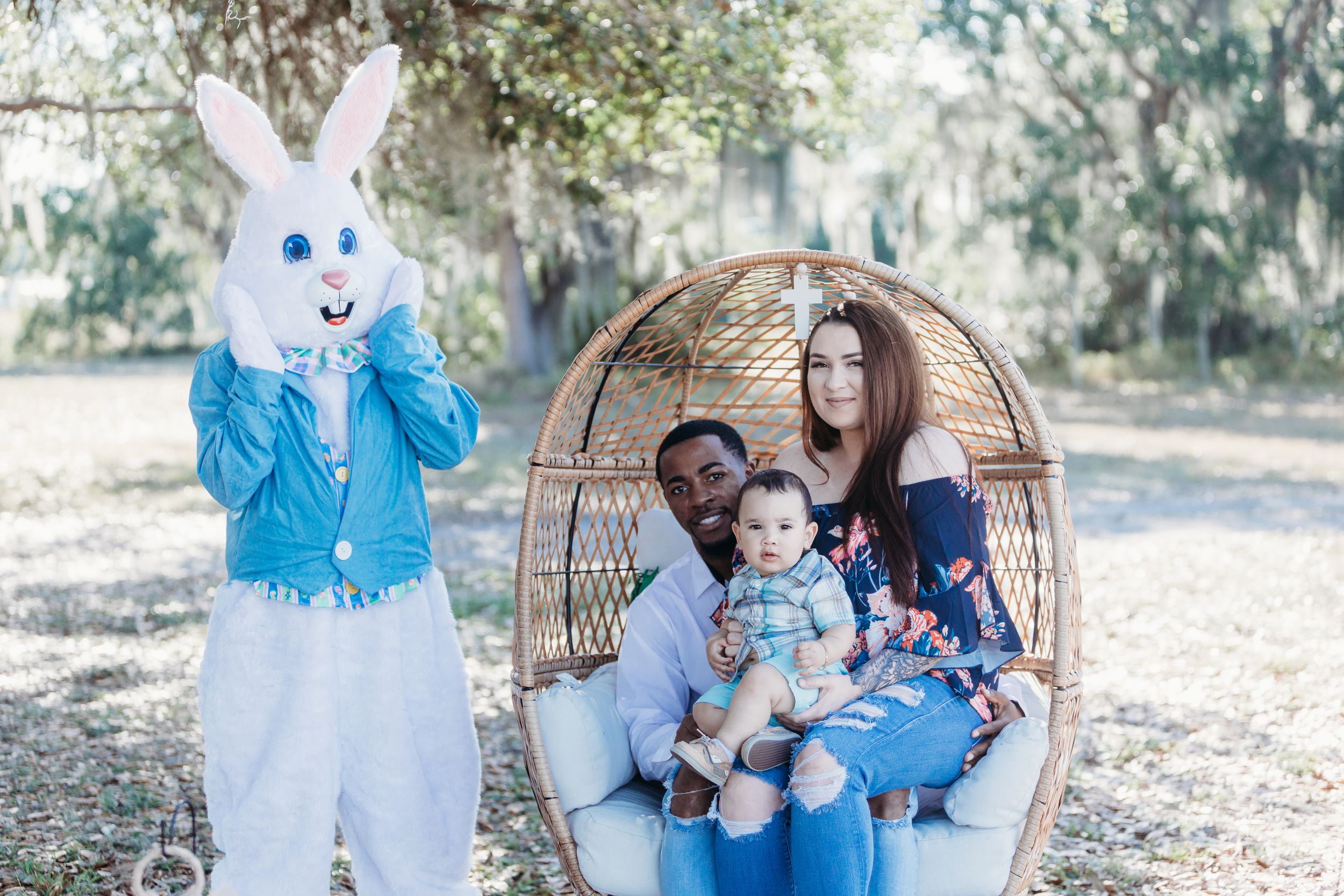 Easter bunny in blue jacket poses next to a wicker egg chair in an outdoor spring setting with moss-draped trees.