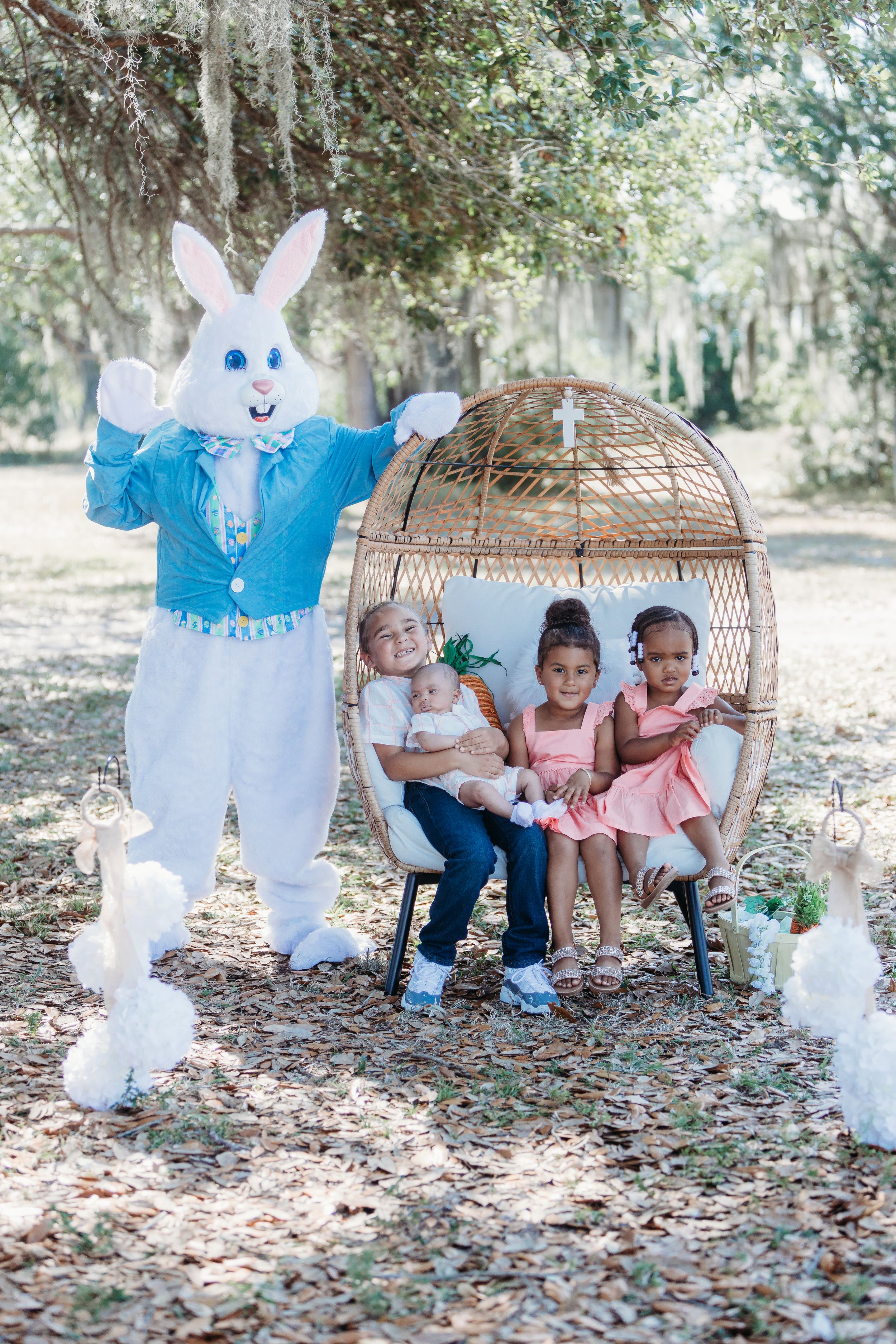 Easter Bunny mascot poses with children sitting in a wicker egg chair outdoors.