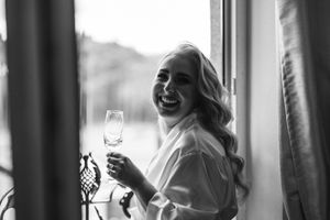 A candid black and white photo of someone laughing while holding a wine glass by a window.