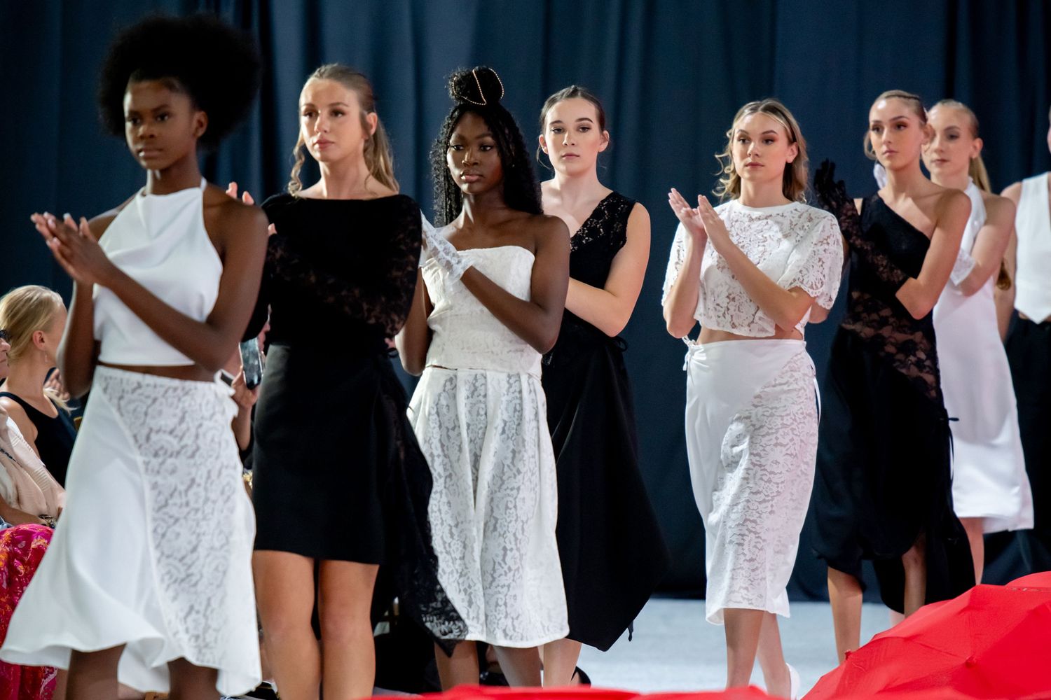 Models in alternating black and white dresses applaud during fashion show finale.