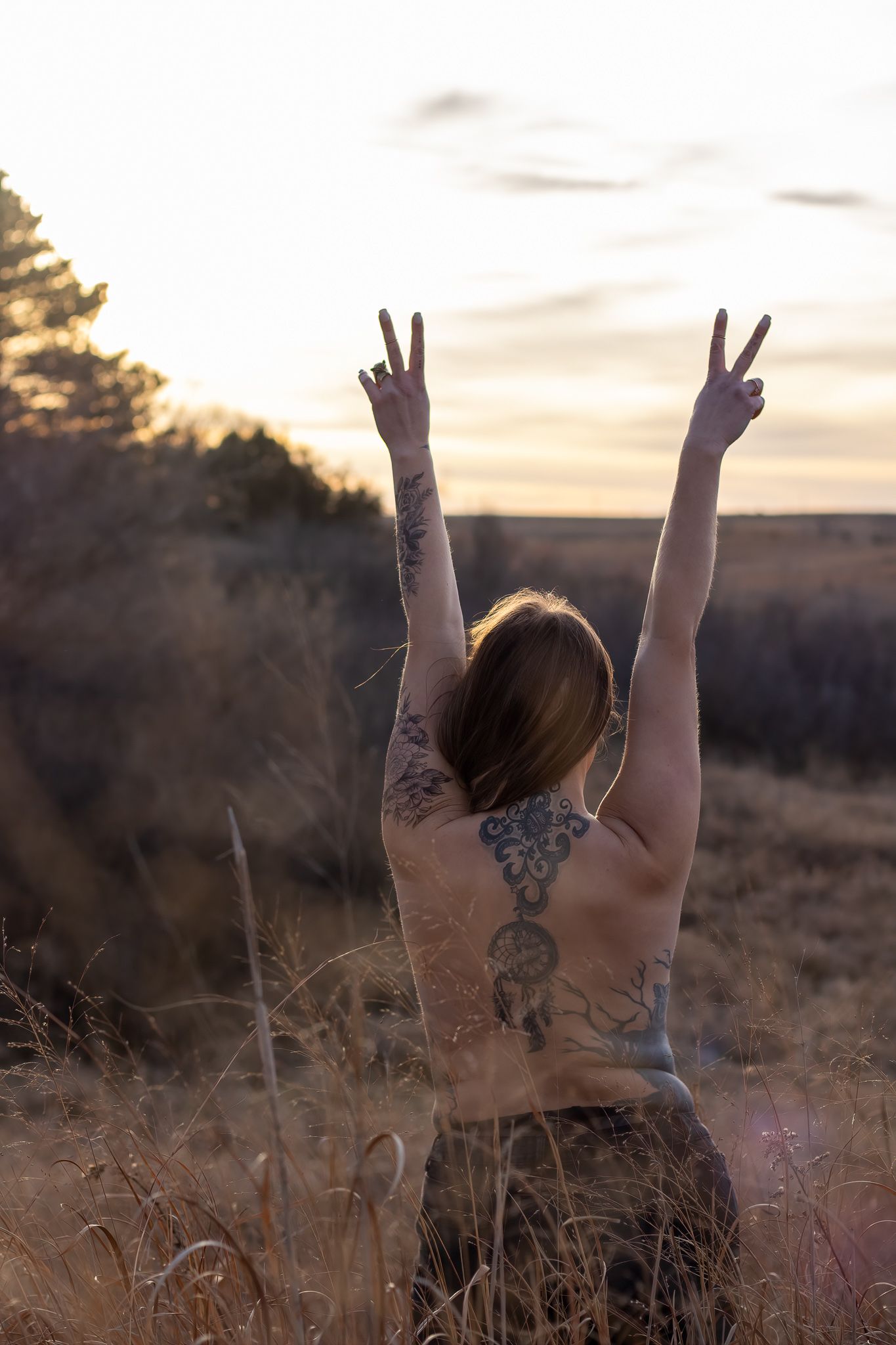 Person with tattoos raising peace signs against golden sunset in field.