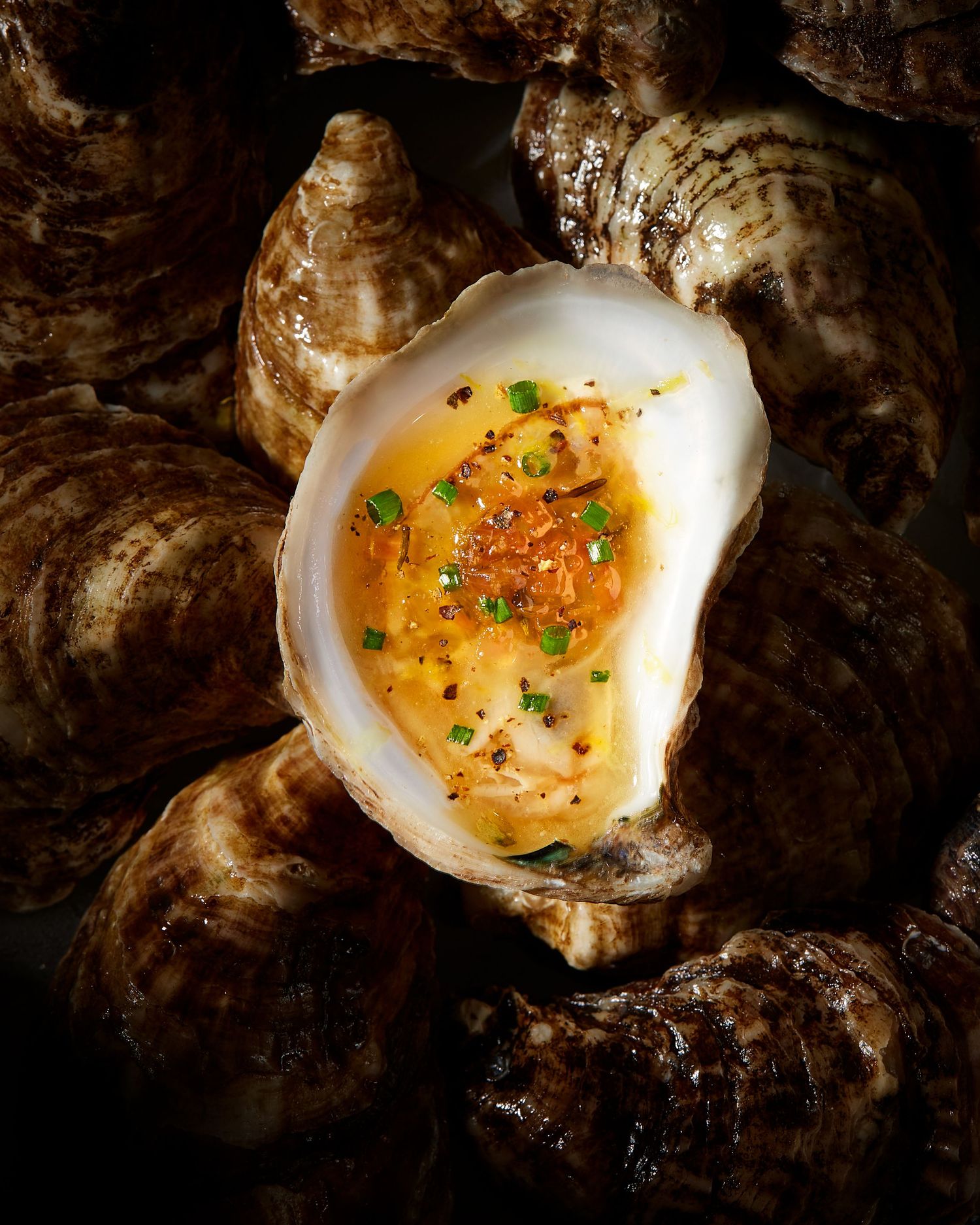 A freshly shucked oyster garnished with citrus oil and fresh herbs sits on a dark rustic stone surface.