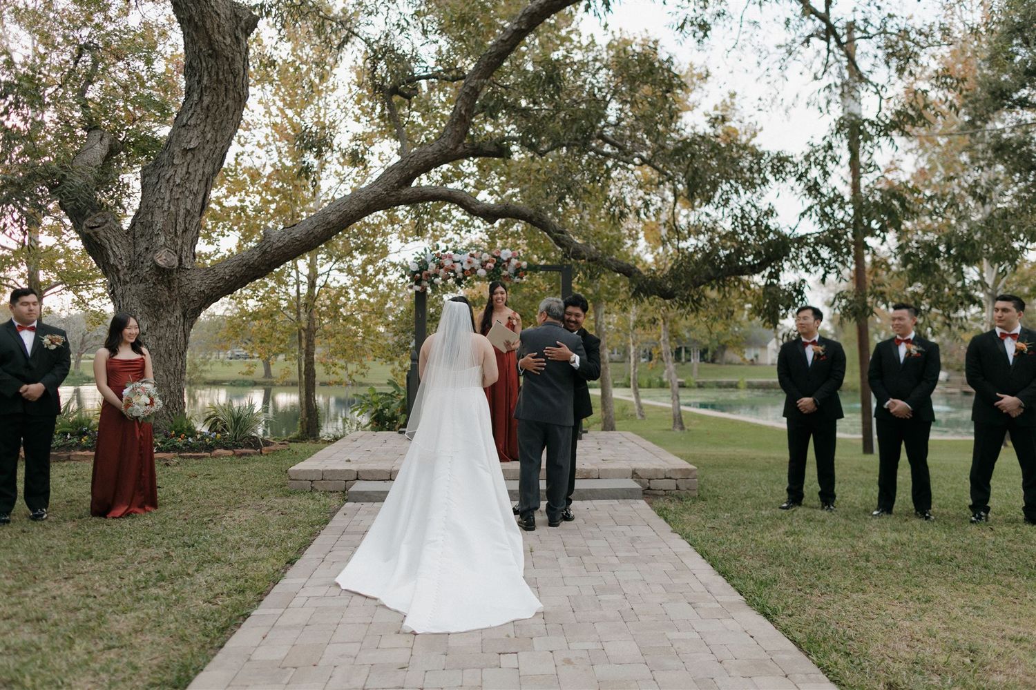 An outdoor wedding ceremony with attendants standing on a stone path under trees.