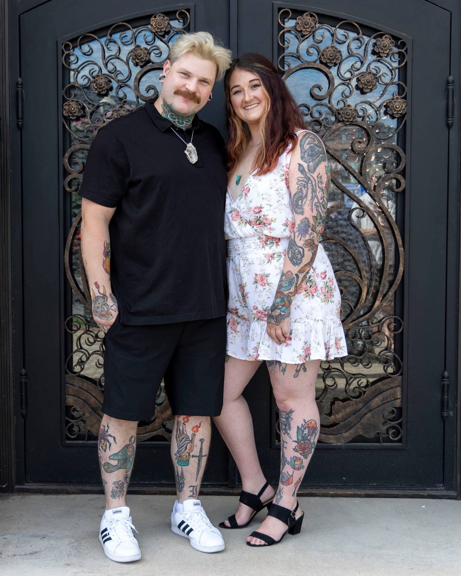 Couple with tattoos poses together against ornate black metal door background.