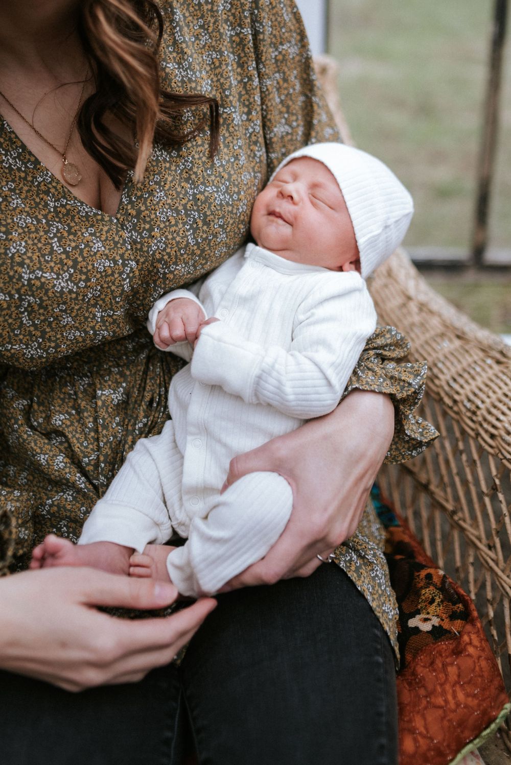 Newborn baby wearing white knit outfit rests peacefully in floral-dressed caregiver's arms.