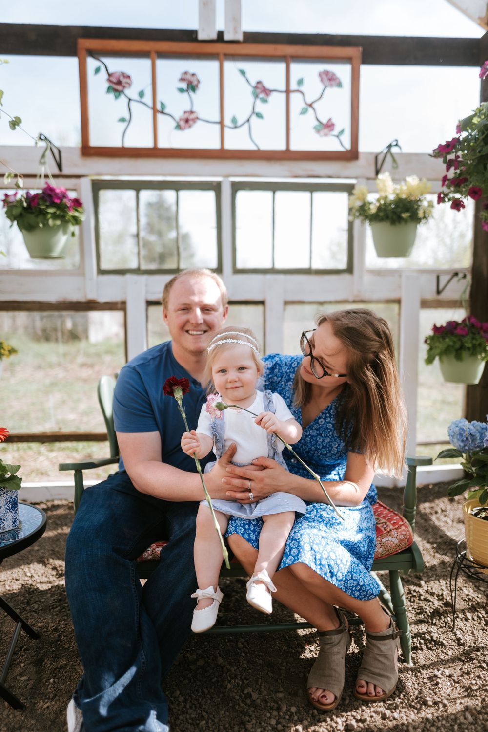 Joyful family portrait in a sunny greenhouse surrounded by potted flowers and rustic decor.