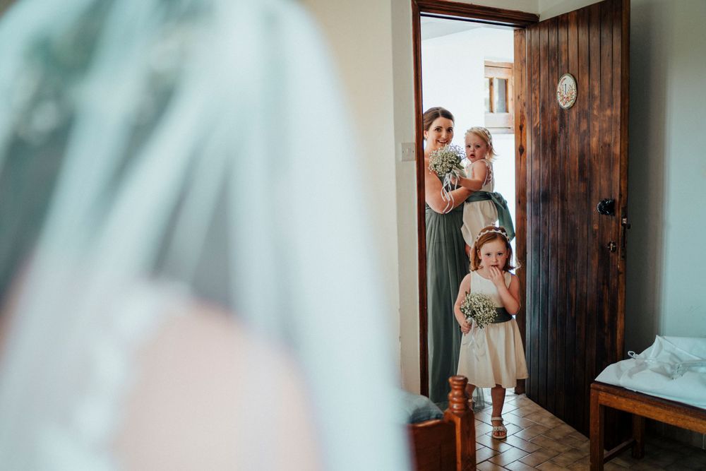 Children peek excitedly through a wooden doorway at a bride in her wedding gown.