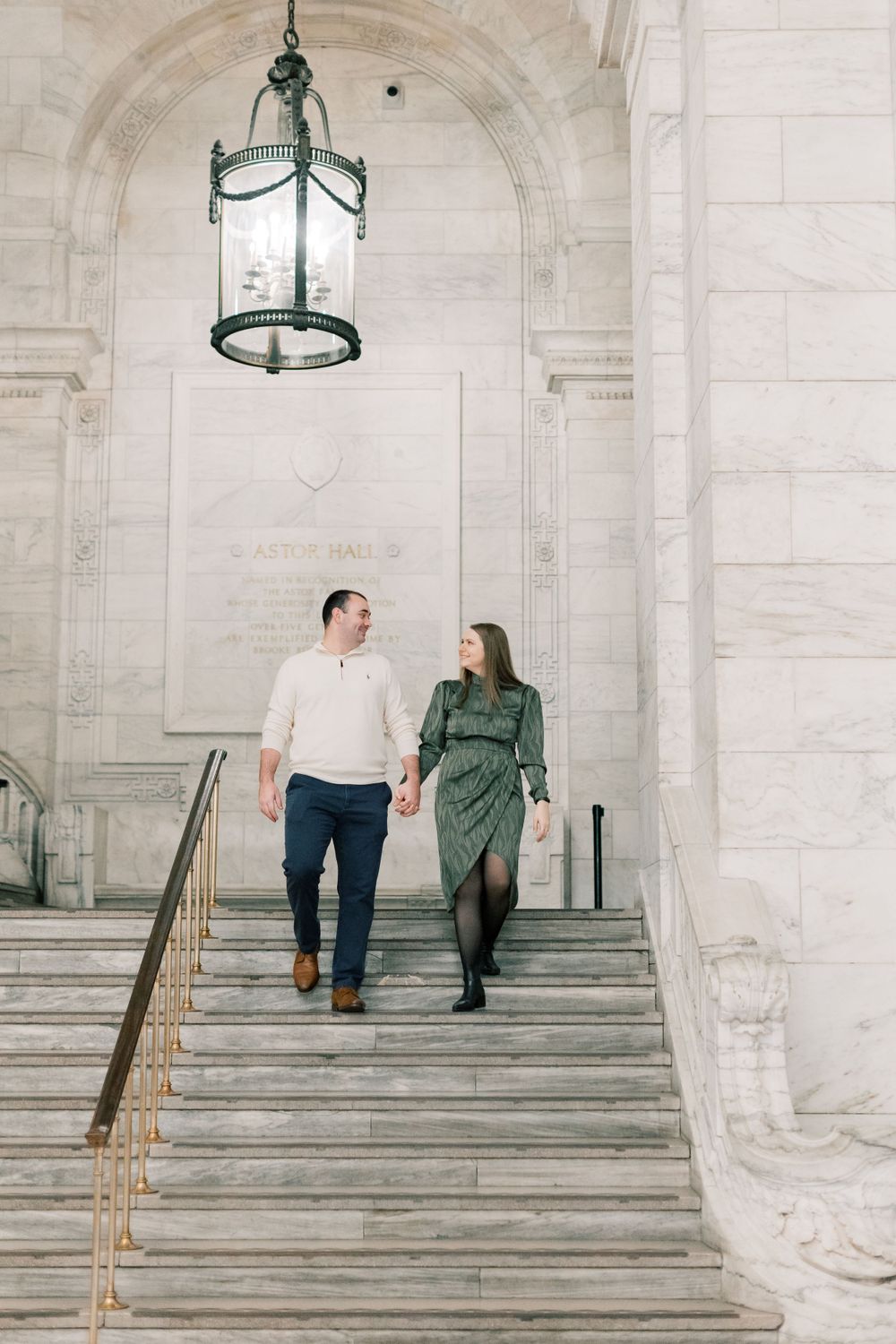 Couple descends grand marble stairs under vintage hanging lantern in historic building.