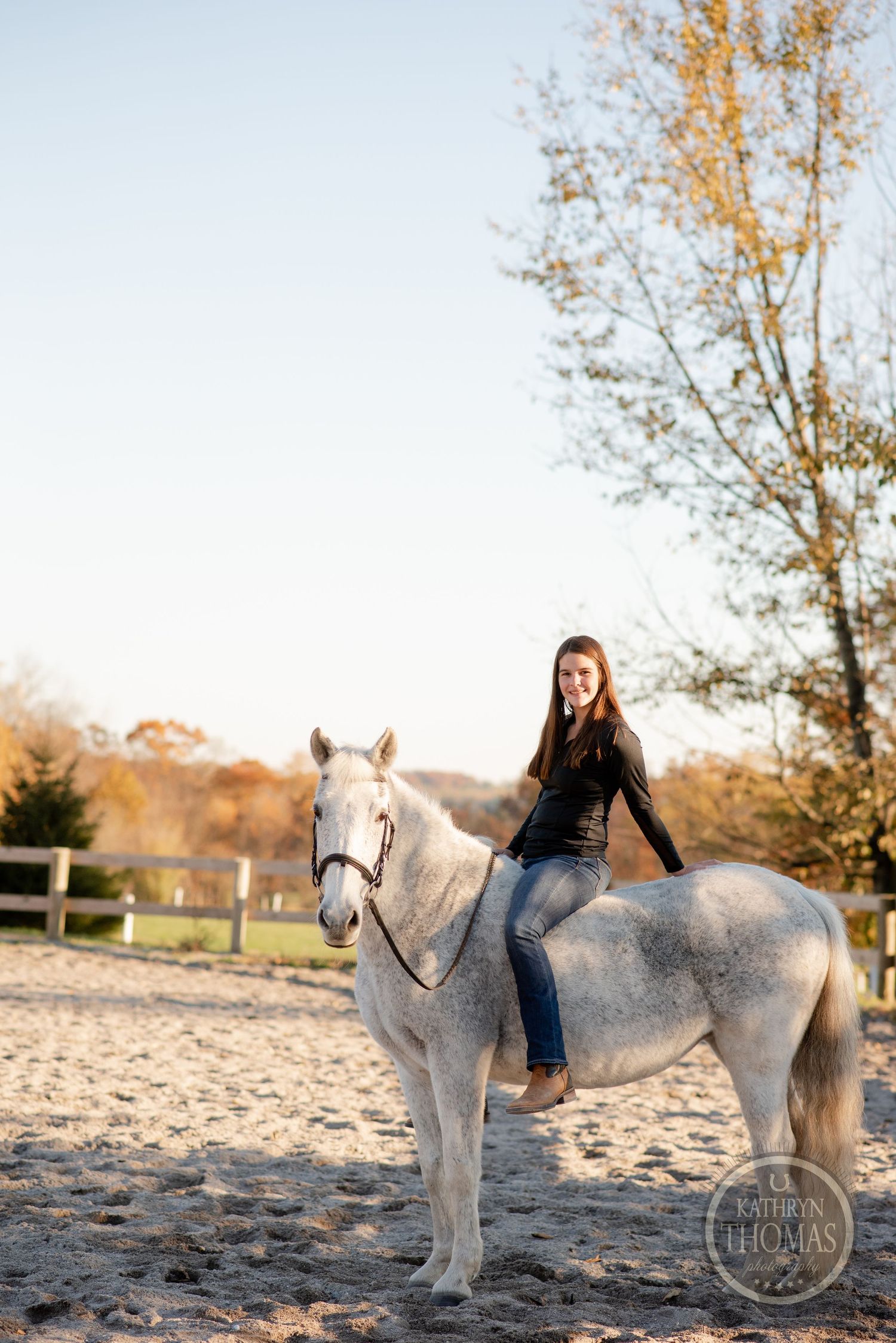 Person riding bareback on a white horse in outdoor arena during autumn sunset.