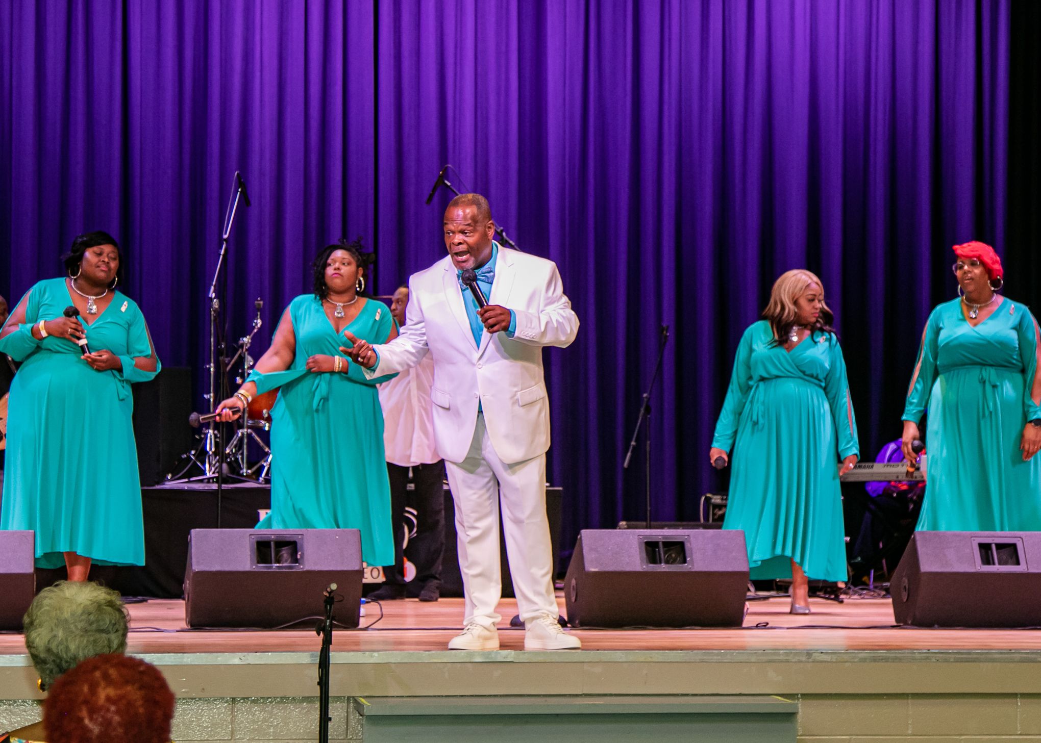Gospel performers in turquoise dresses and white suit singing on purple-curtained stage.