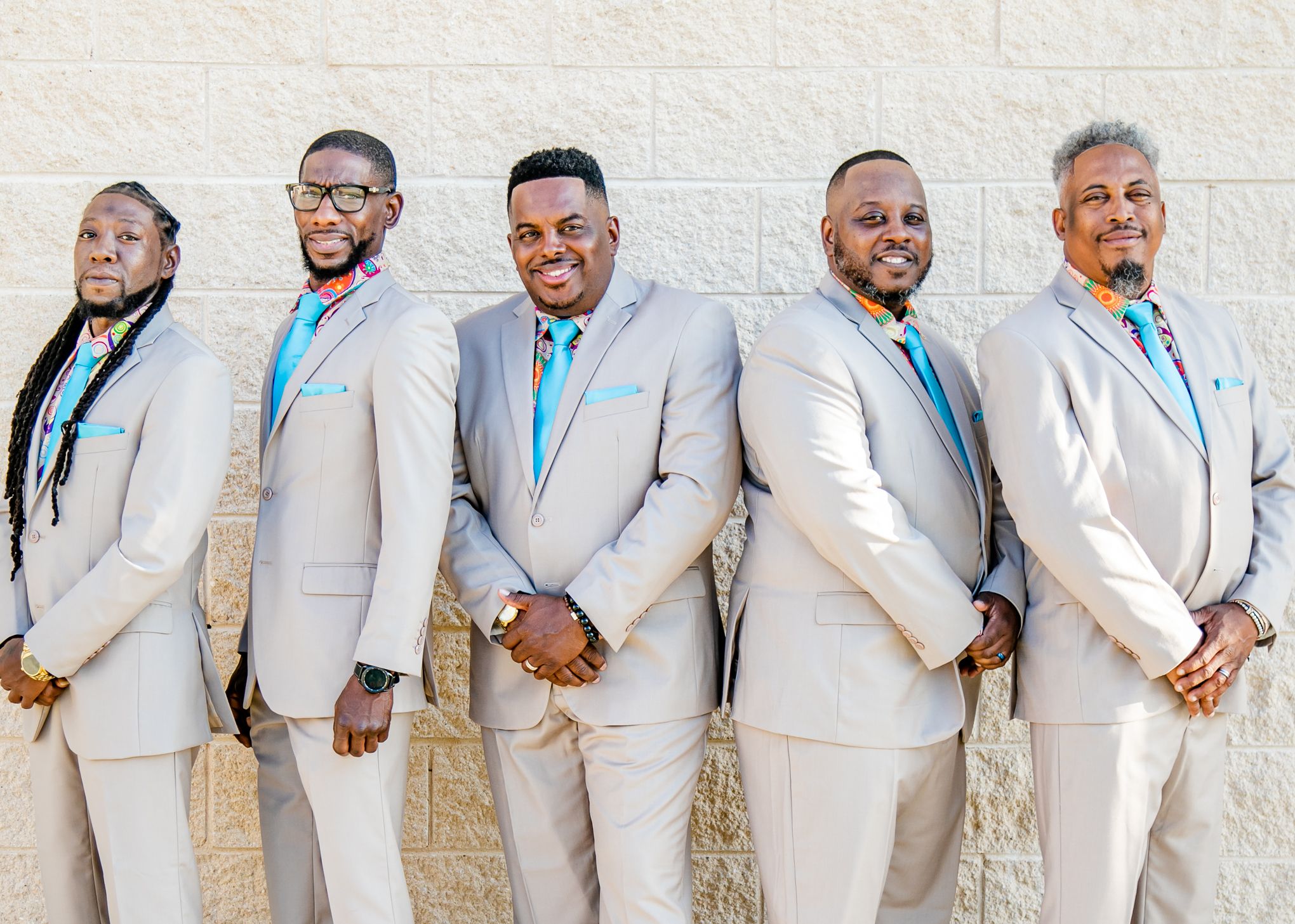 Groomsmen in light gray suits with blue ties pose against stone wall.
