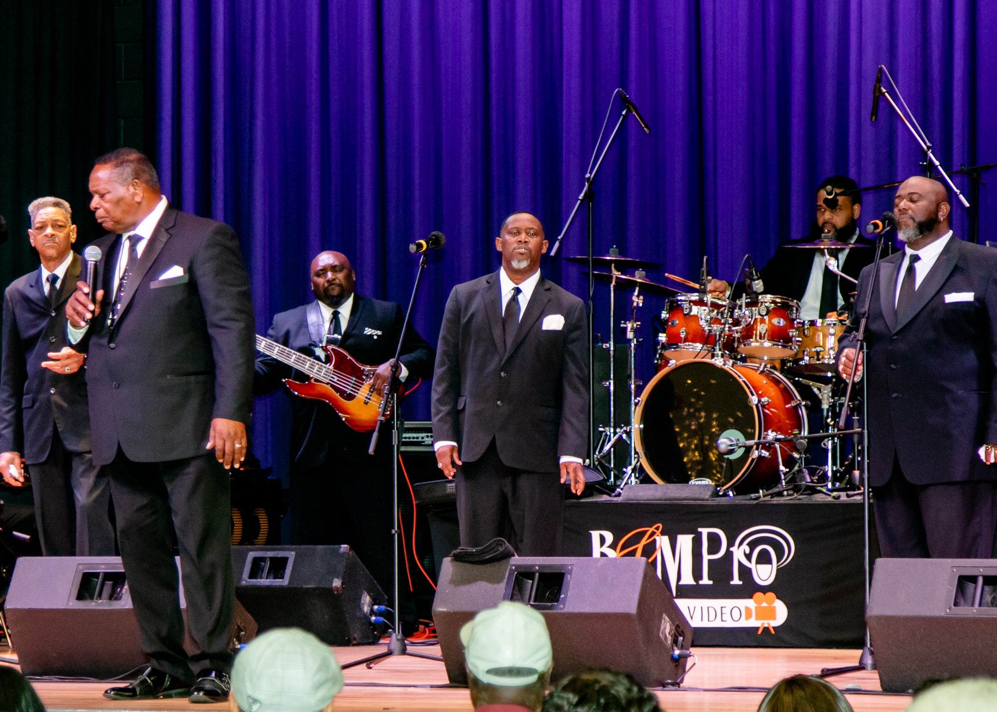 Musical group performs on stage with instruments and purple curtain backdrop.
