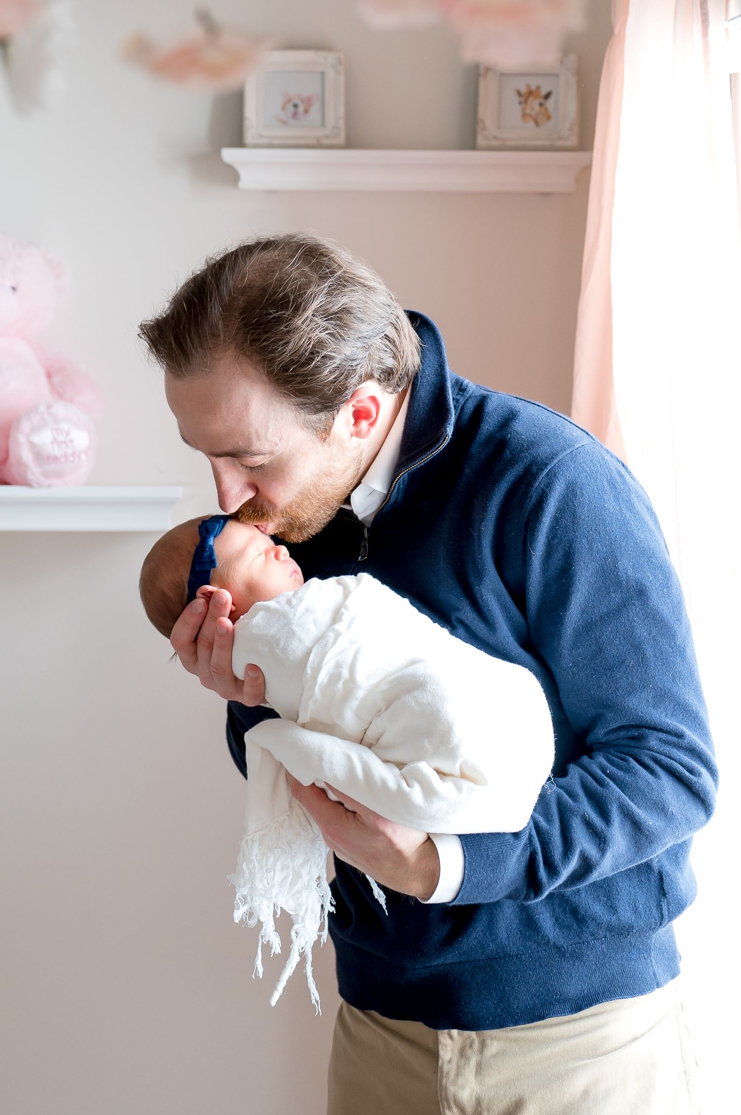 father wearing a blue sweater kissing the forehead of his newborn baby girl inside her pink soft nursery in west u