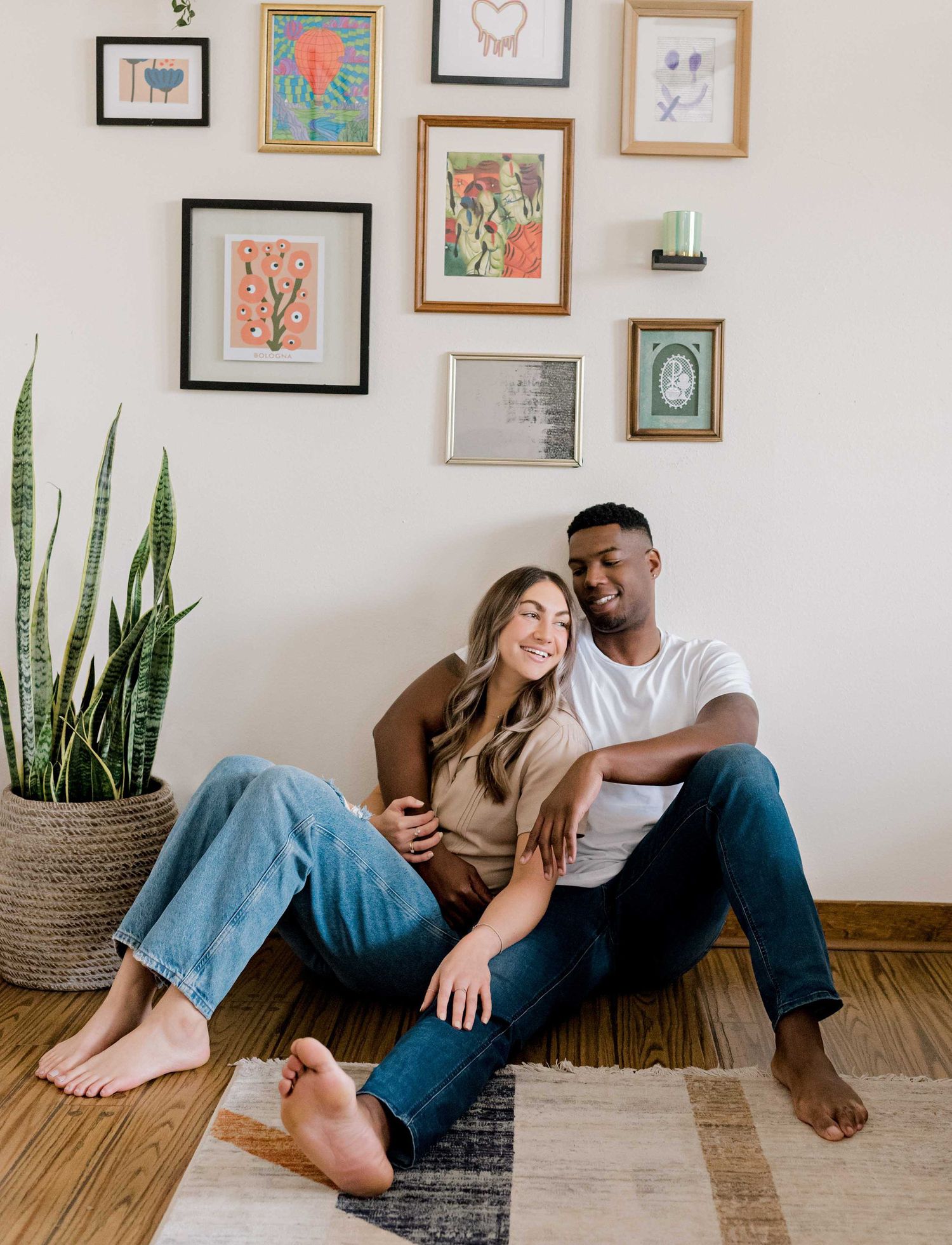 Couple sitting on hardwood floor beneath gallery wall with snake plant decor.