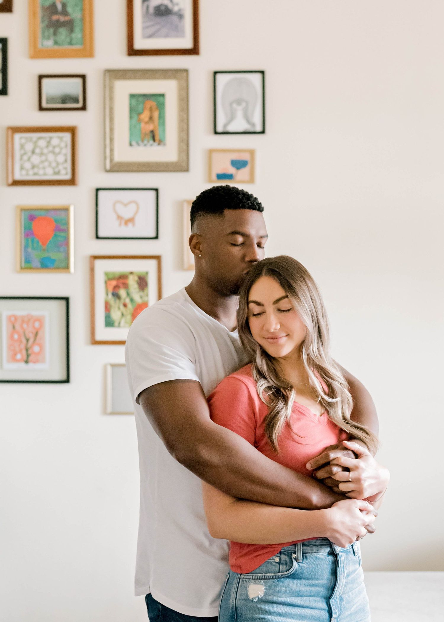 A tender embrace in front of a gallery wall with framed artwork in a bright minimalist room.