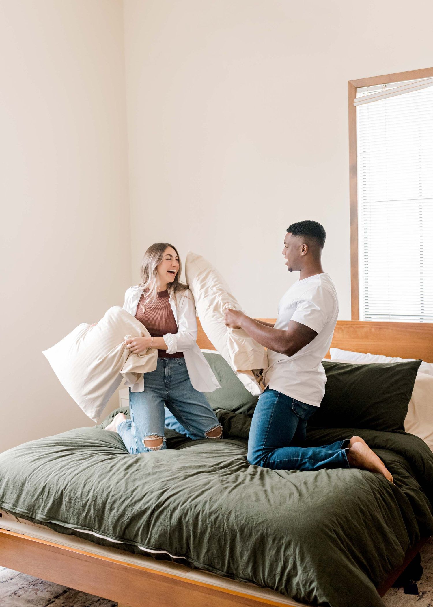 Playful pillow fight on a bed with olive green bedding in a bright bedroom.