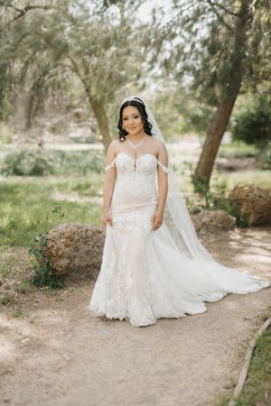 Bride in lace wedding gown with long train standing on nature path.