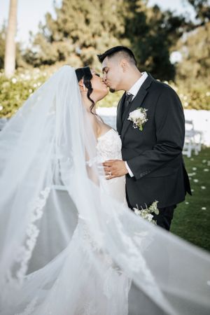 Wedding couple sharing kiss with flowing white veil in outdoor garden setting.