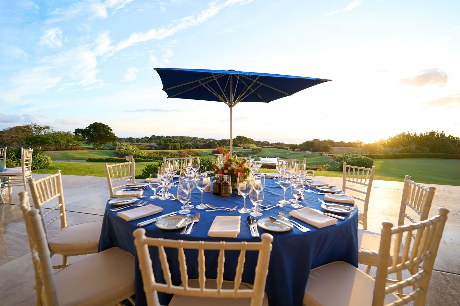 Elegant outdoor dining table with blue linens and umbrella set against sunset view.