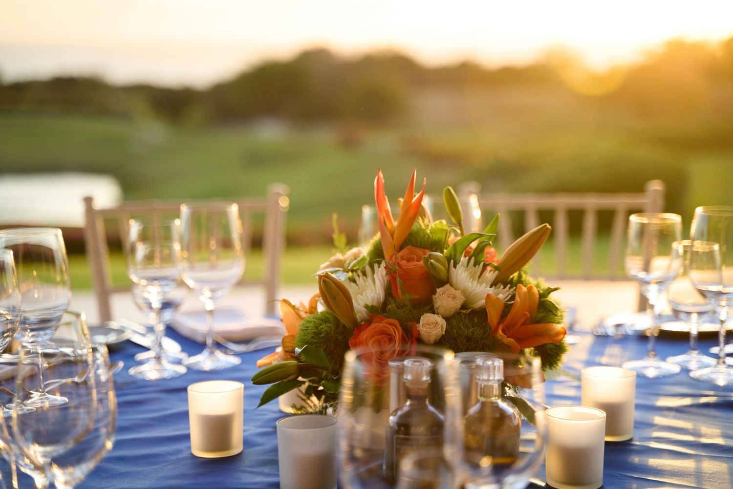 Table centerpiece with orange flowers and candles on blue tablecloth at sunset.