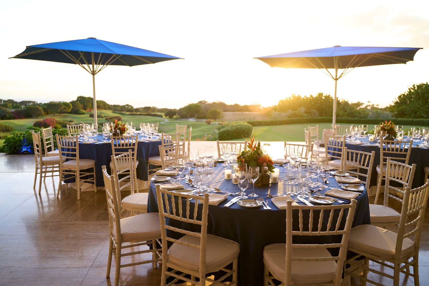 Outdoor golf club dining setup with blue tablecloths and umbrellas against sunset golf course view.